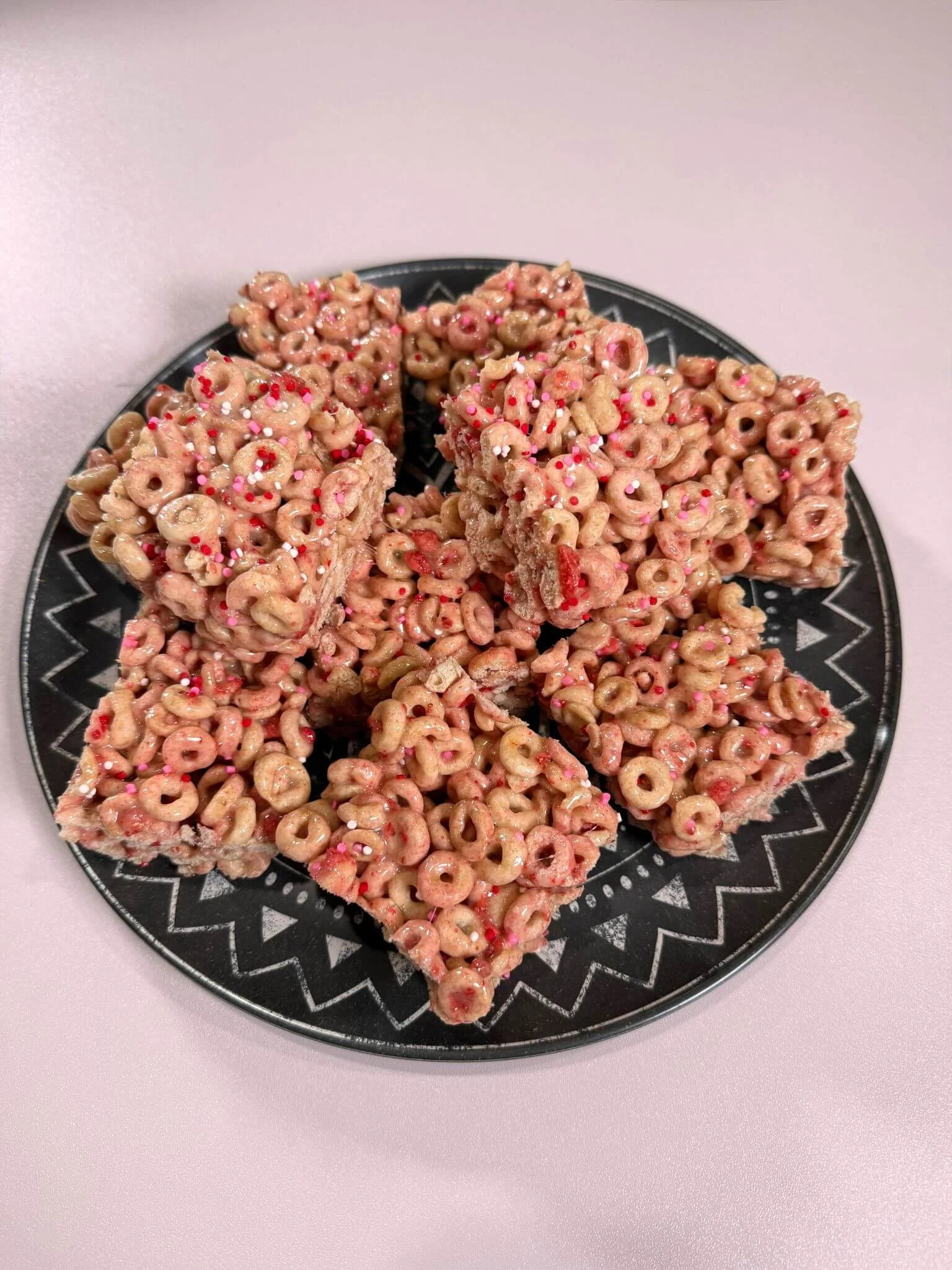 a white table with a black printed plate full of cereal bar looking desserts. desserts are pink and have sprinkles.