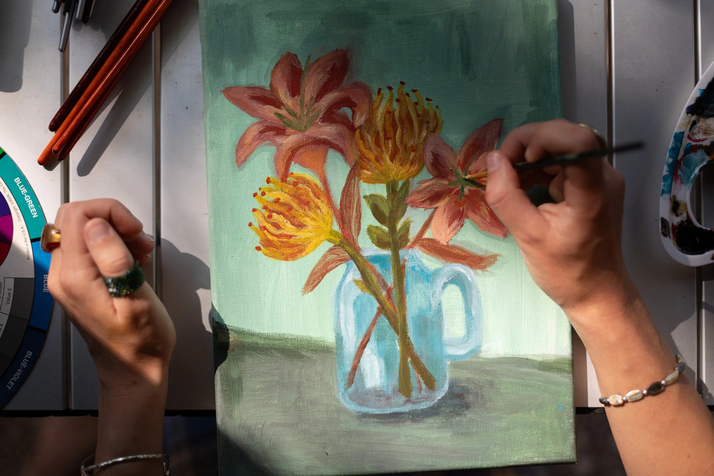 Person painting a still life of pink lilies and yellow flowers in a light blue mug on a canvas with oil paints and brushes.