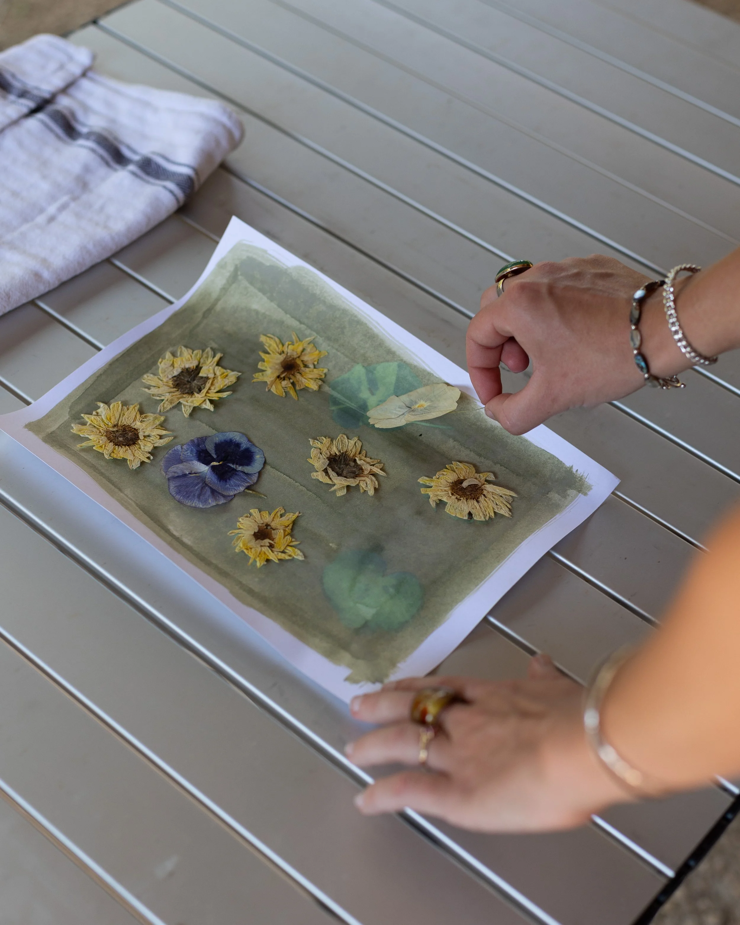 Person arranging pressed flowers on paper with a towel nearby on a metal table