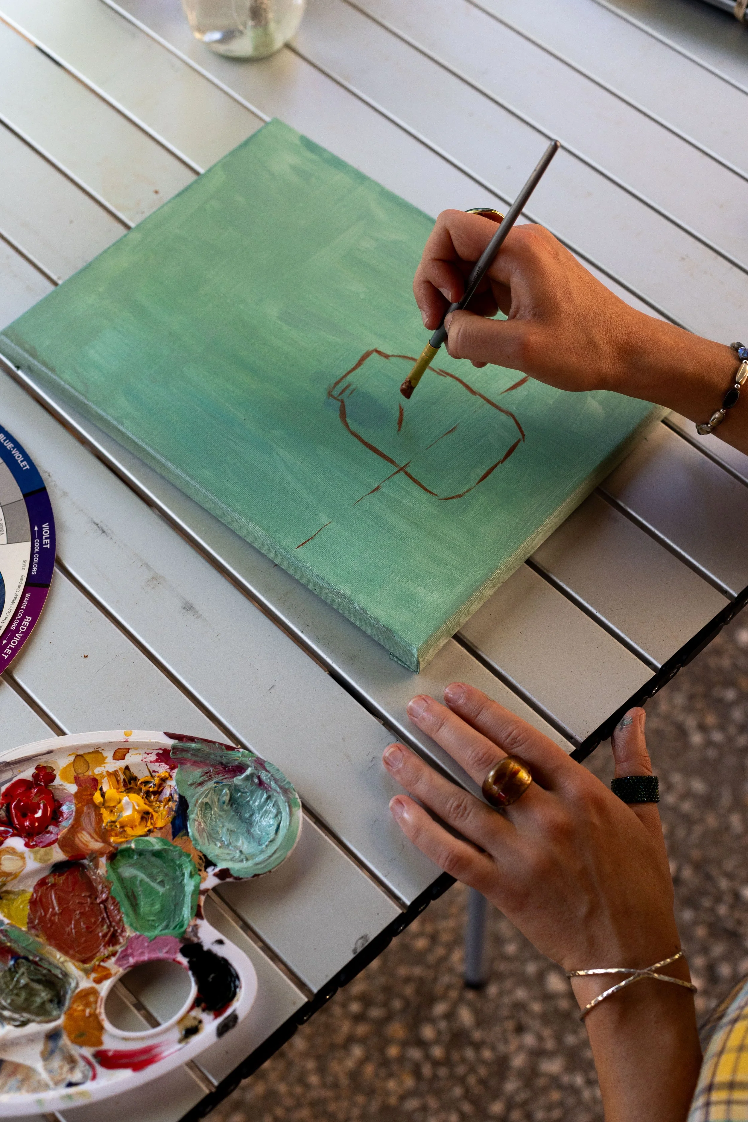Person painting a landscape on a canvas with a palette of colors nearby, seated at a white outdoor table.