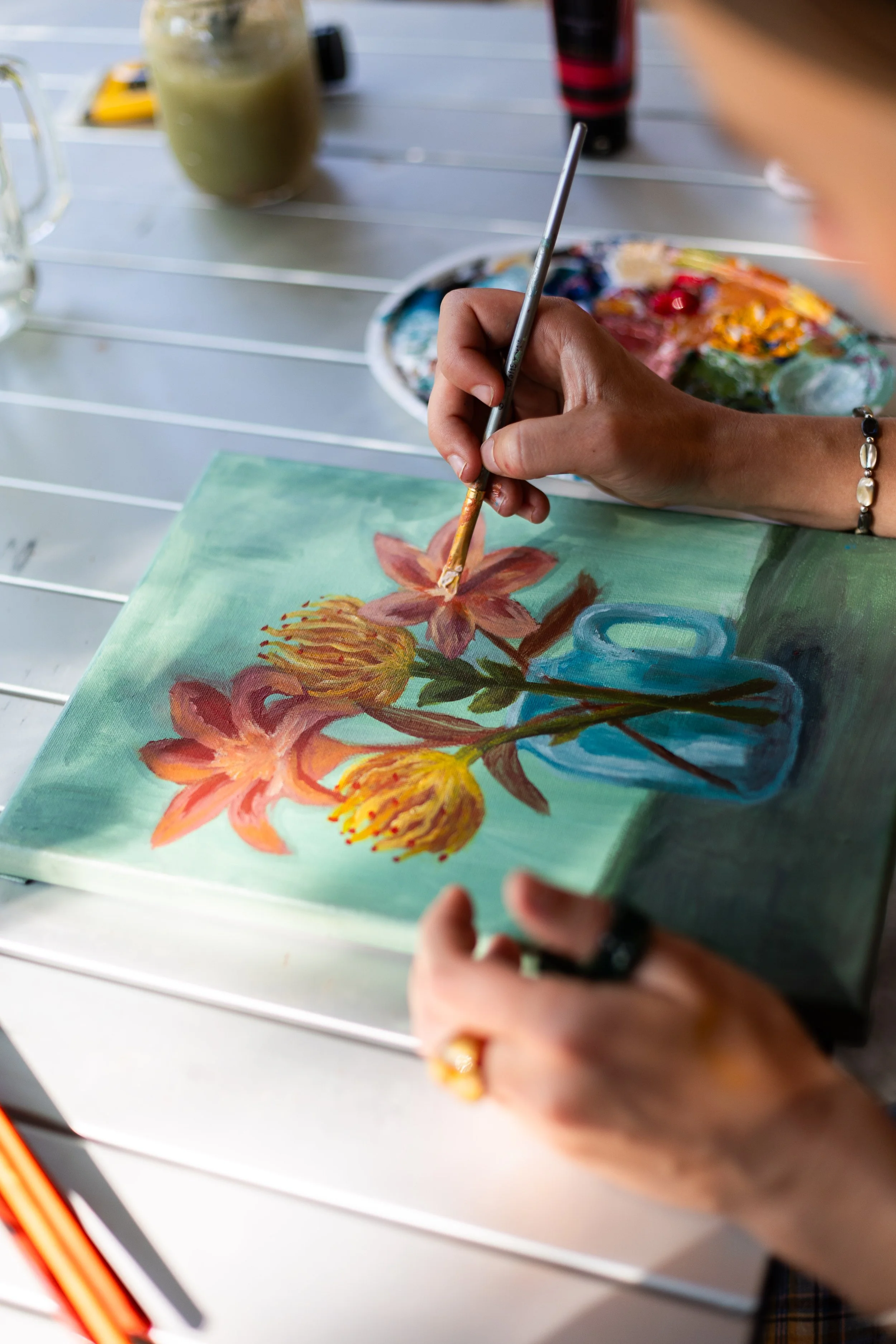 Person painting a floral still life on canvas, with a palette of colors nearby, on a white outdoor table.