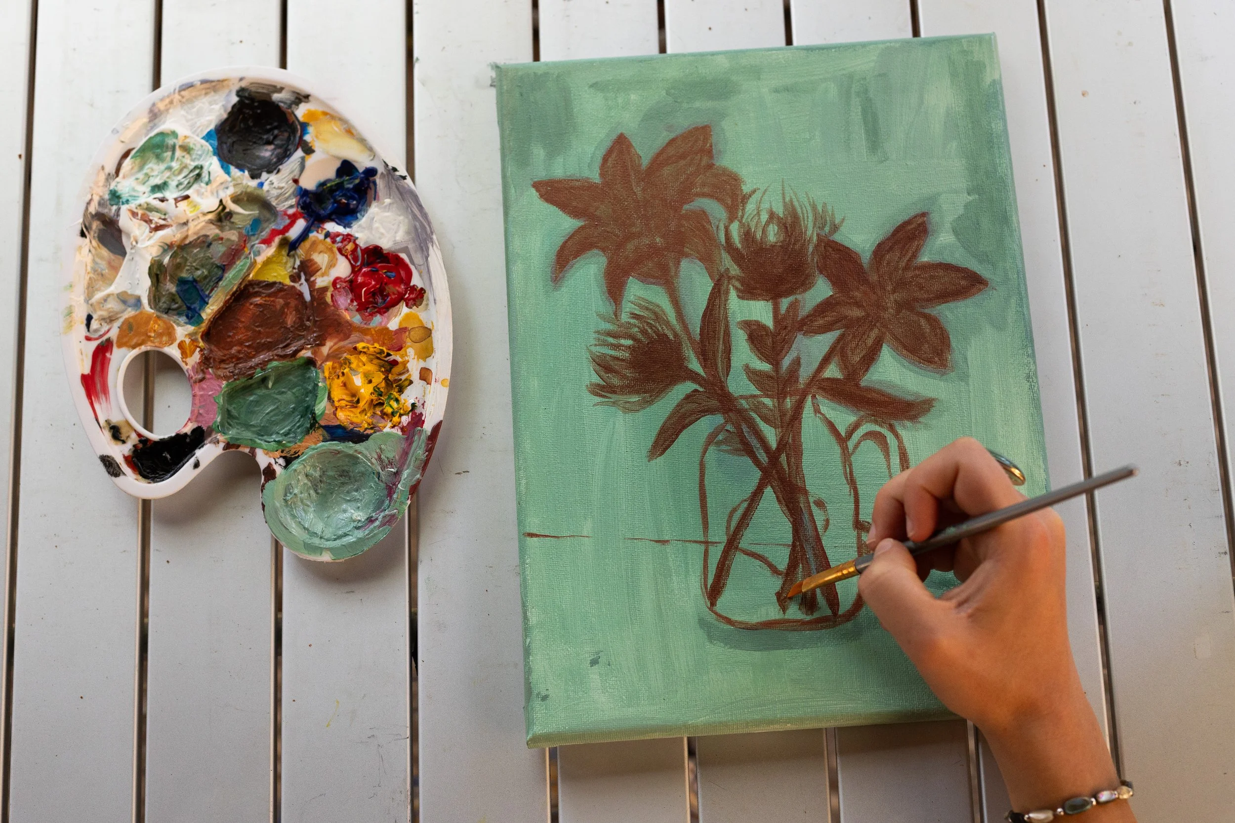 A person painting a floral still life on a canvas, with a palette of mixed colors nearby.