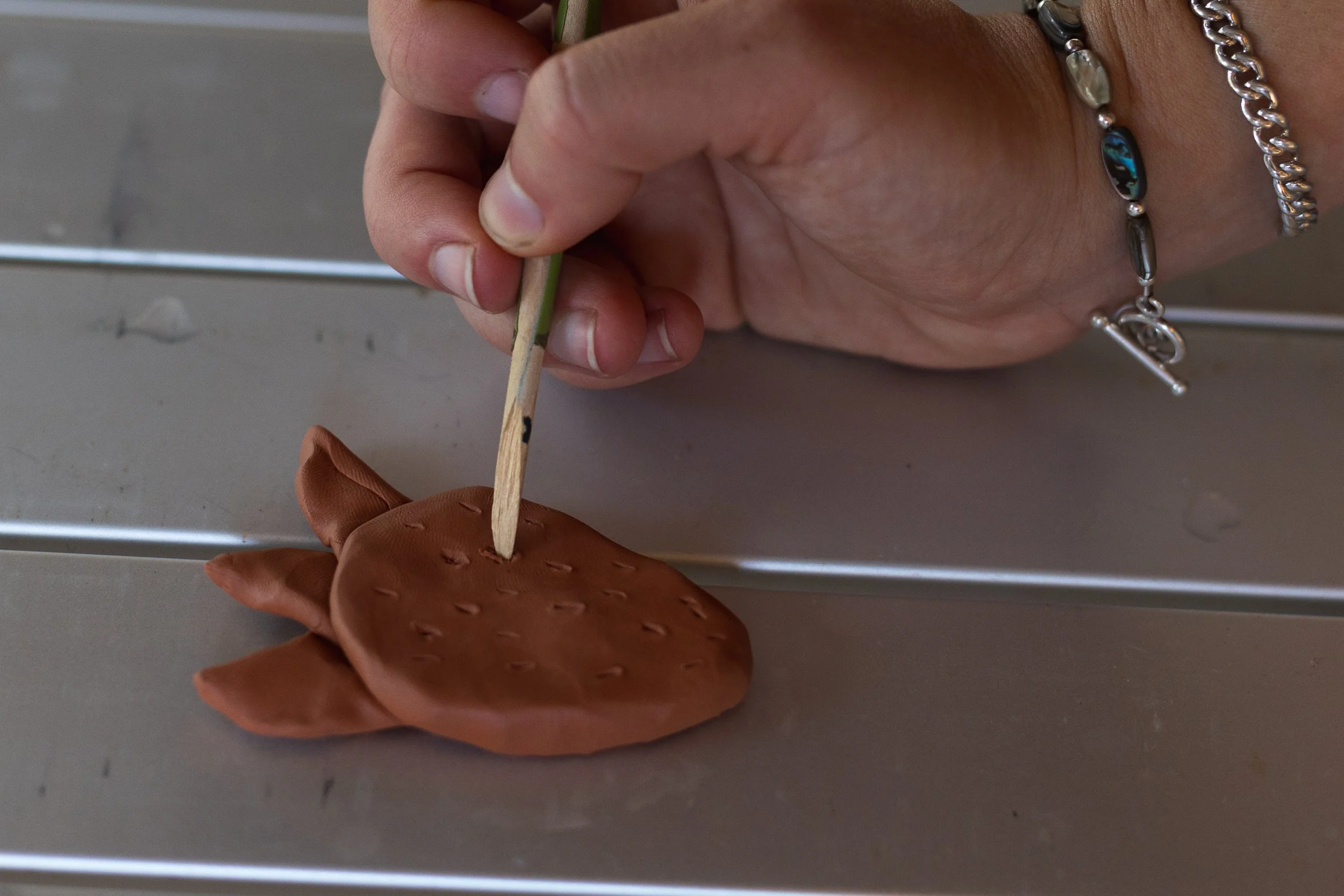 A person's hand is holding a small wooden stick, inserting it into a clay sculpture of a fish. The person is wearing a bracelet and a chain bracelet on their wrist. The scene appears to be on a metallic surface.