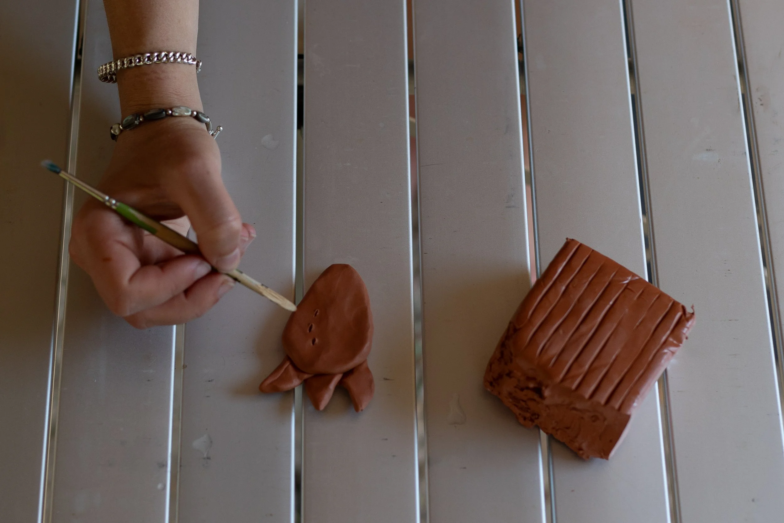 Hand painting details on a small clay fish sculpture with a paintbrush, next to a larger clay fish sculpture on a metallic surface.