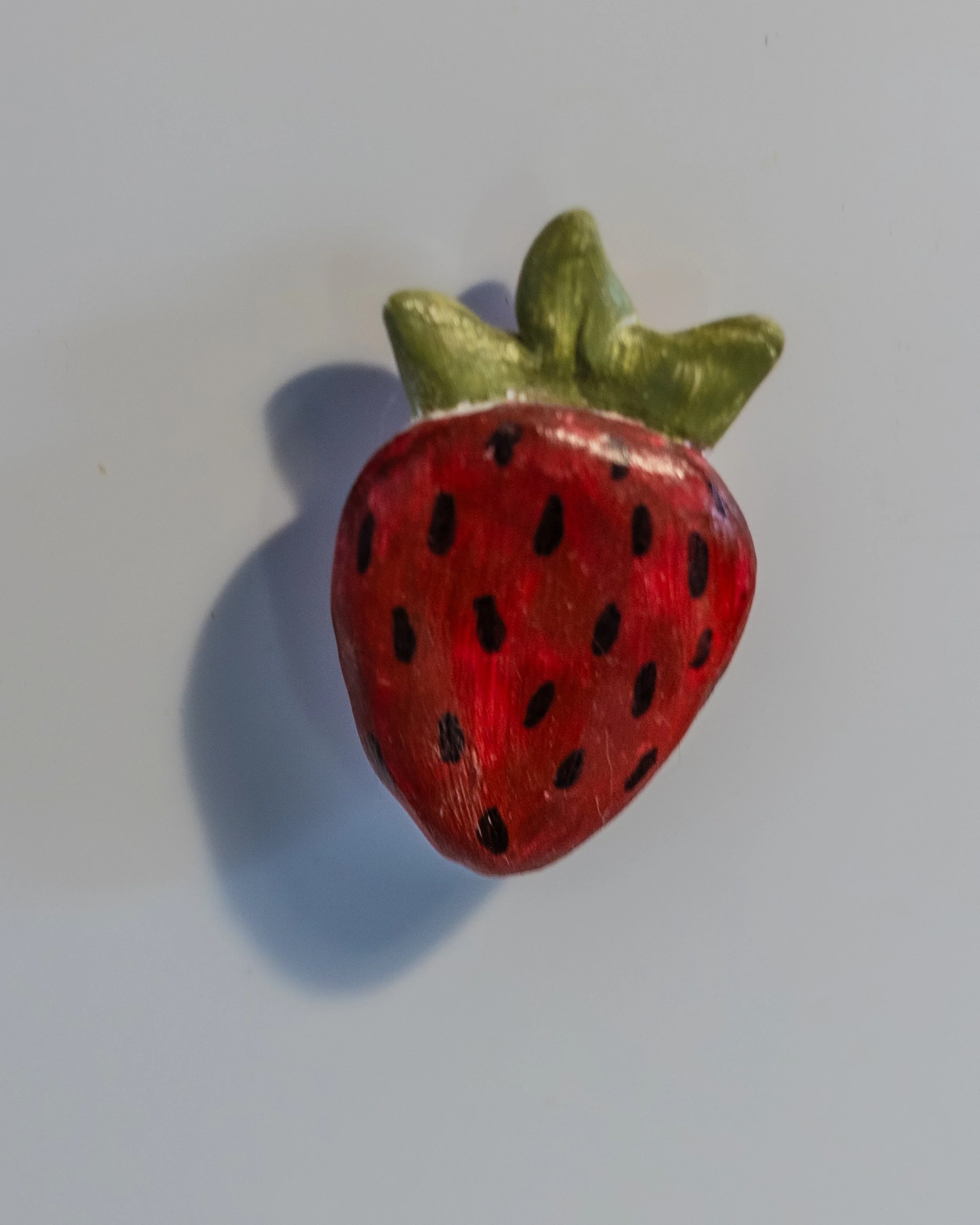A small, realistic-looking strawberry figurine with green leaves on top, cast a shadow on a white surface.
