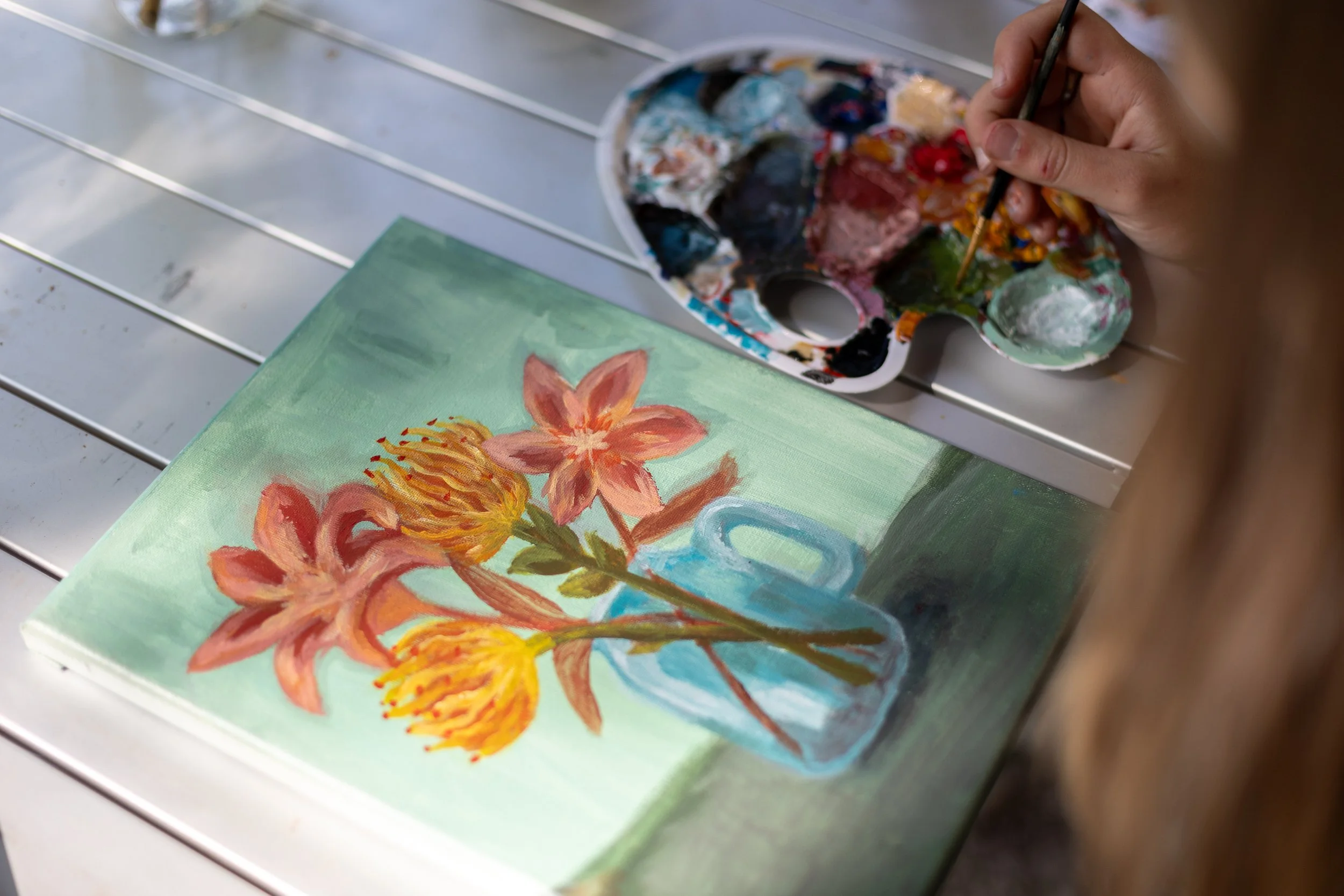 Person painting a still life of pink and yellow flowers in a blue vase on a green background.