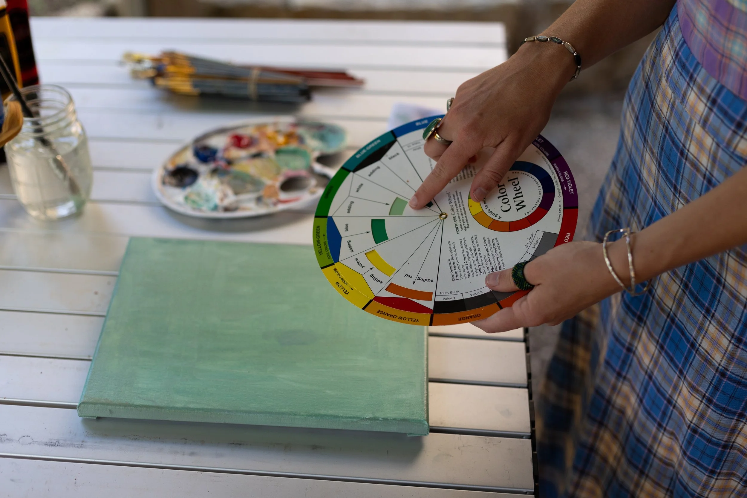 A person holding a color wheel next to a green canvas and an artist's palette on a white table outdoors.