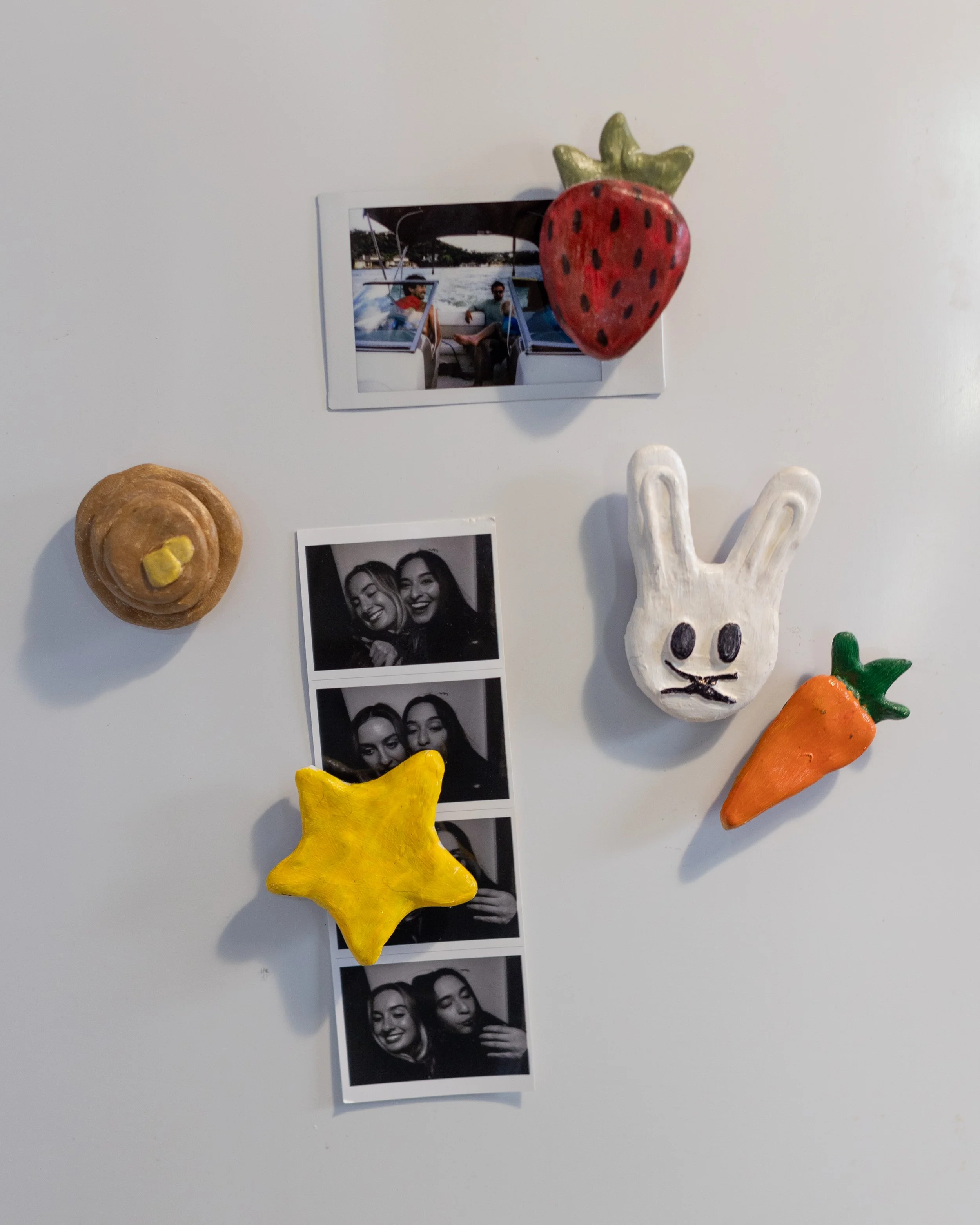 White wall decorated with a photo, a photo booth strip, a strawberry magnet, a cinnamon roll magnet, a bunny magnet, and a carrot magnet.