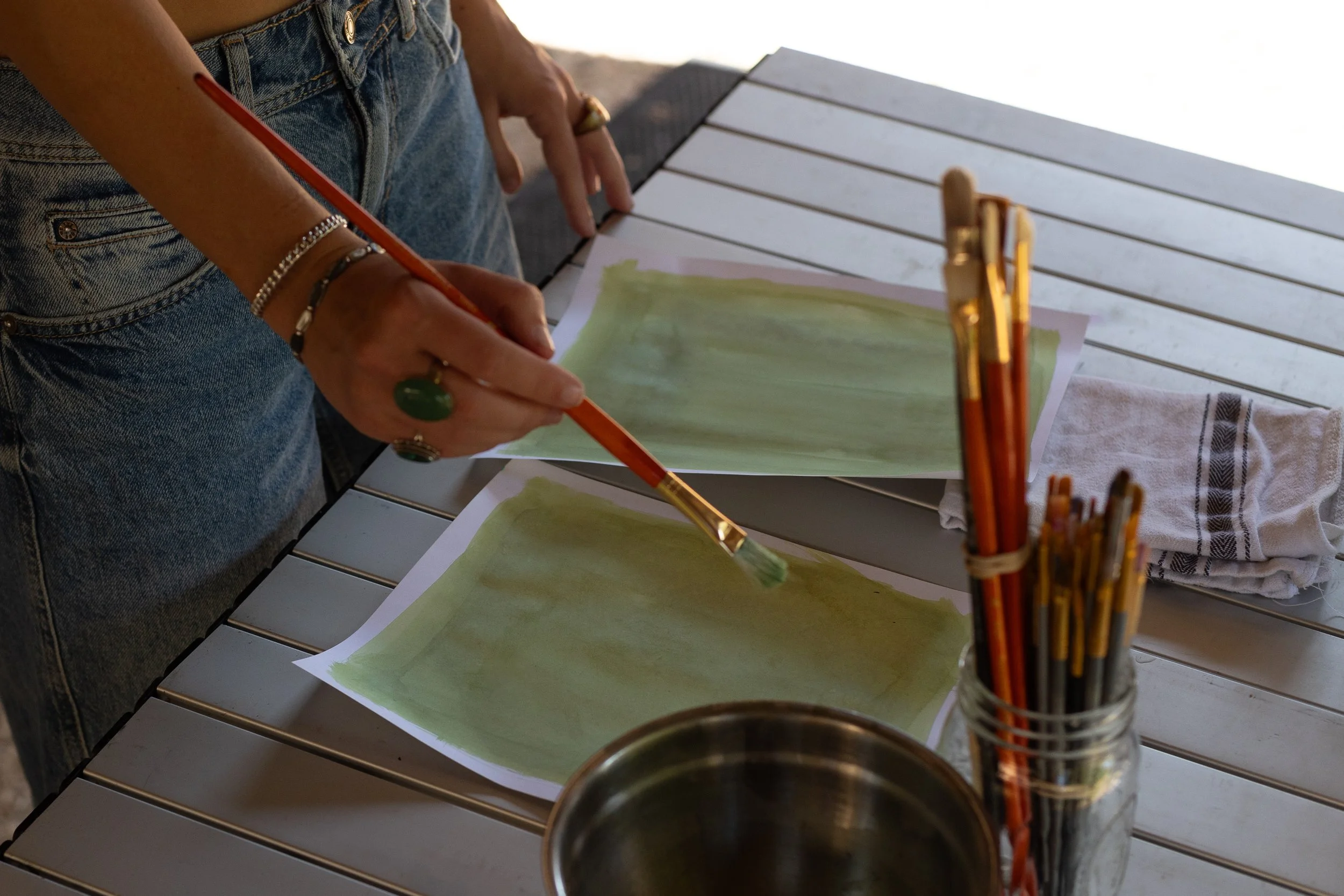Person painting with a green watercolor brush on paper, with brushes in a jar nearby and a cloth on the table.