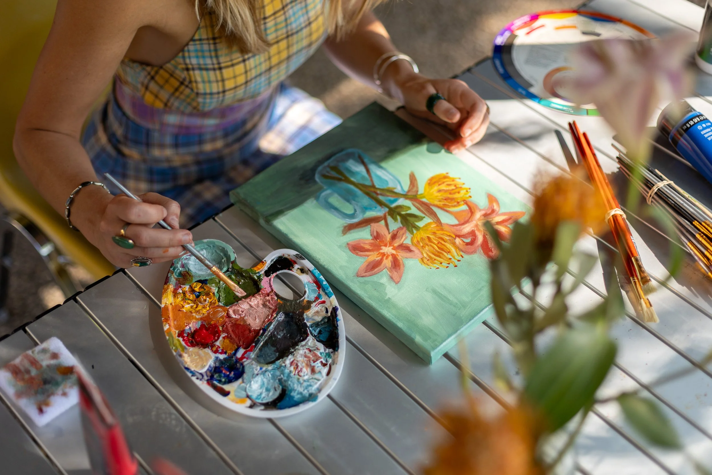 Person painting a floral artwork on a canvas with a palette of paint and brushes on a table, outdoors.