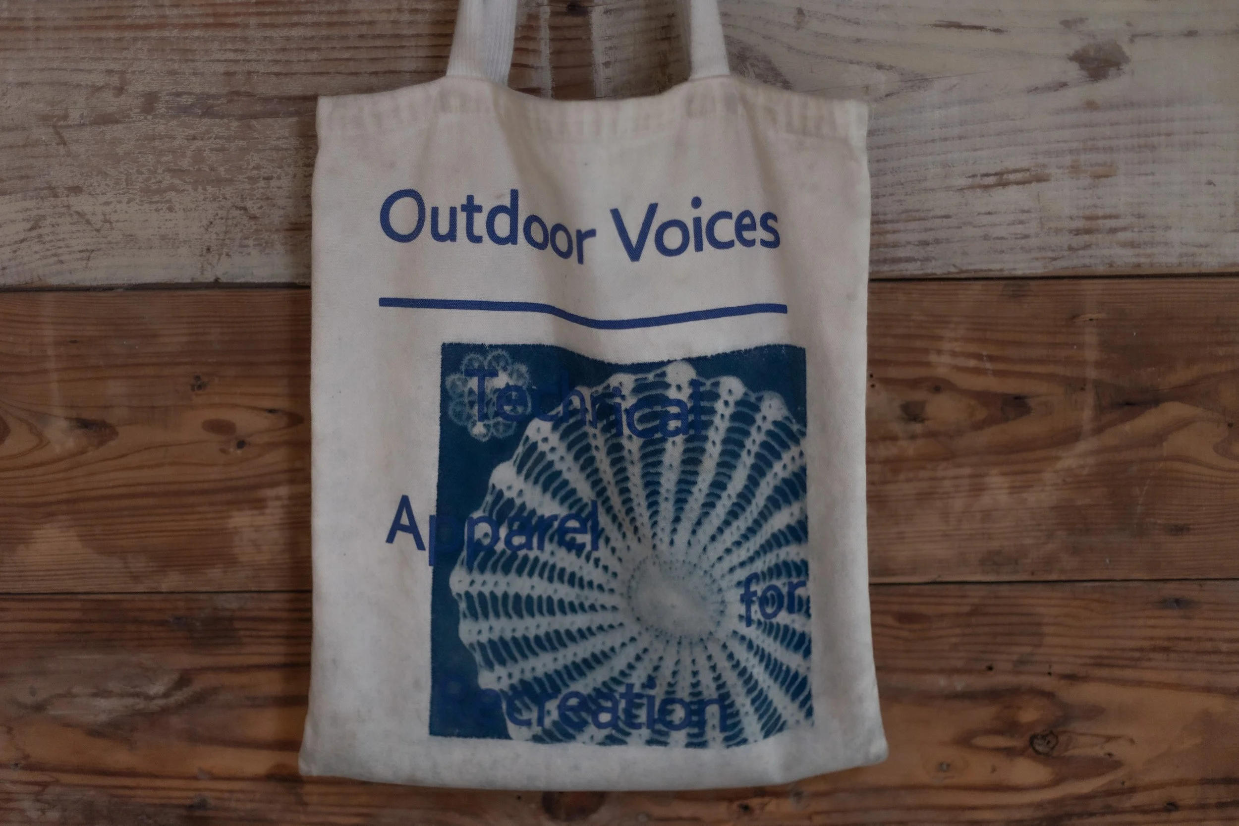 Canvas tote bag with blue text 'Outdoor Voices' and a graphic of a seashell and bubbles, hanging on a wooden wall.
