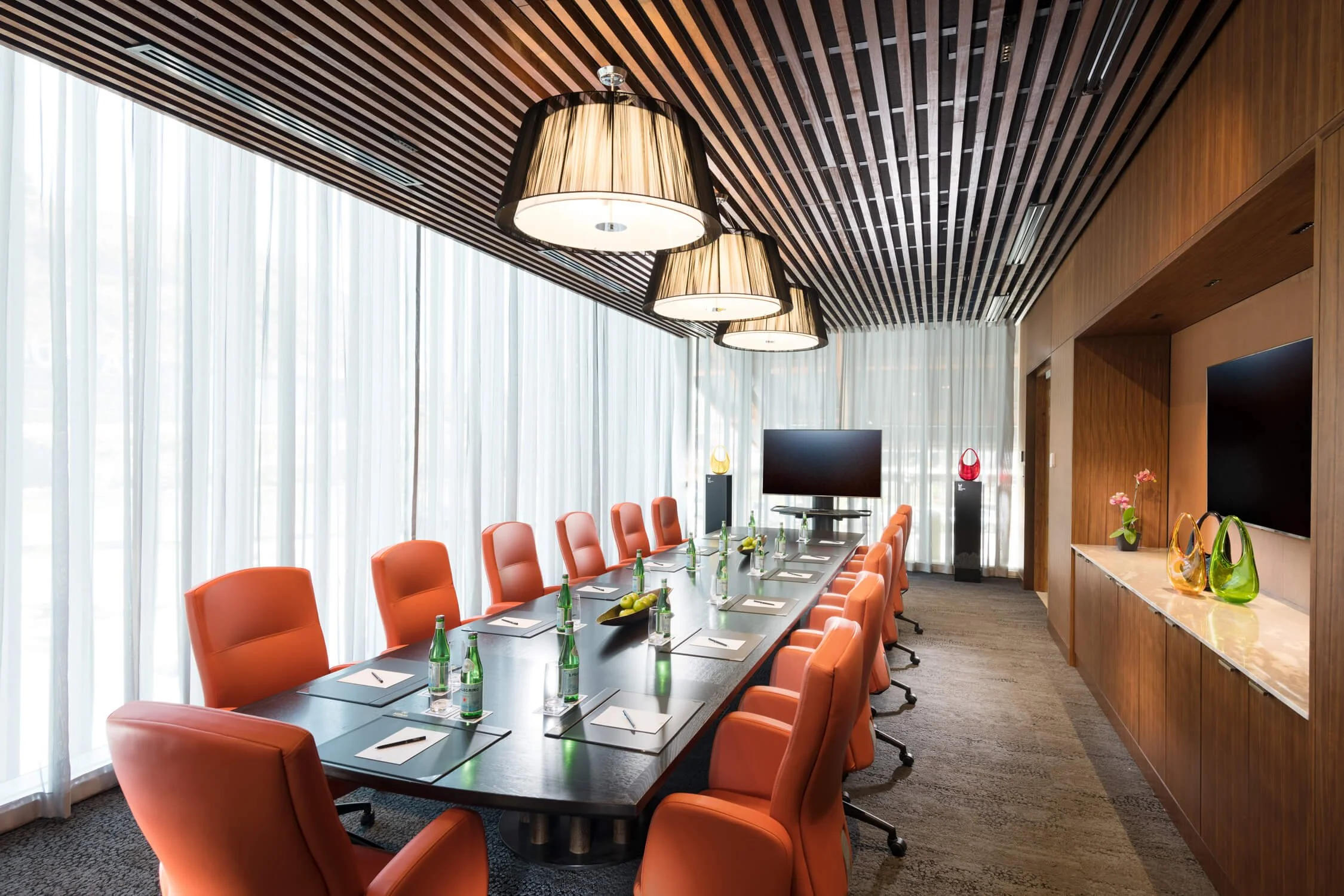 Modern conference room with a long dark wood table, orange chairs, and hanging pendant lights. Large windows with sheer curtains, flat-screen monitors, and decorative vases on a wood-paneled wall.