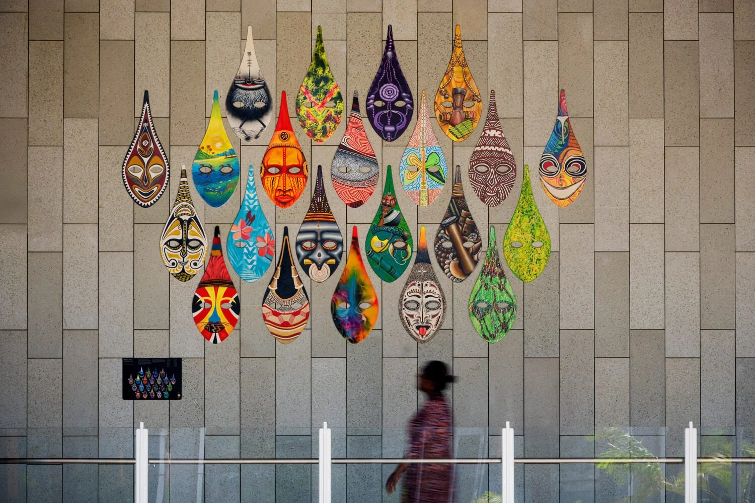 Colorful abstract masks hung on a tiled wall, with a person walking past in a patterned shirt.
