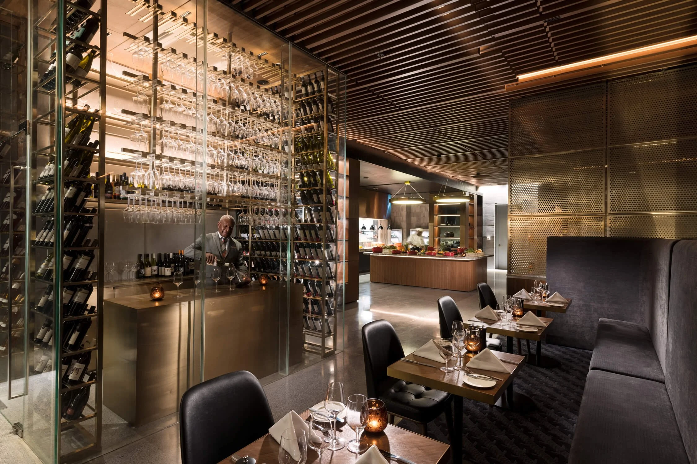 Elegant restaurant dining area with tables set with napkins, glasses, and candles. A wine and glass display behind a bartender preparing drinks, and a buffet station in the background. Modern lighting and decor.
