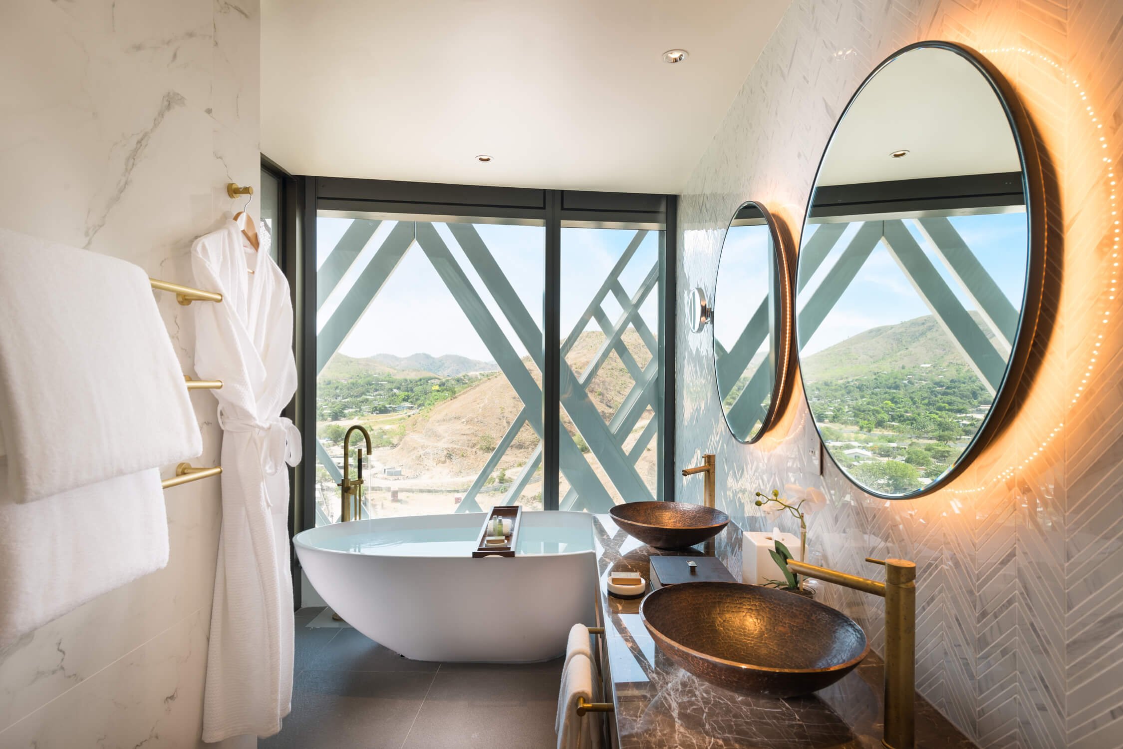 Modern bathroom with a freestanding bathtub, two round mirrors with backlit lighting, gold fixtures, and a view of mountains through large glass windows with geometric patterns.