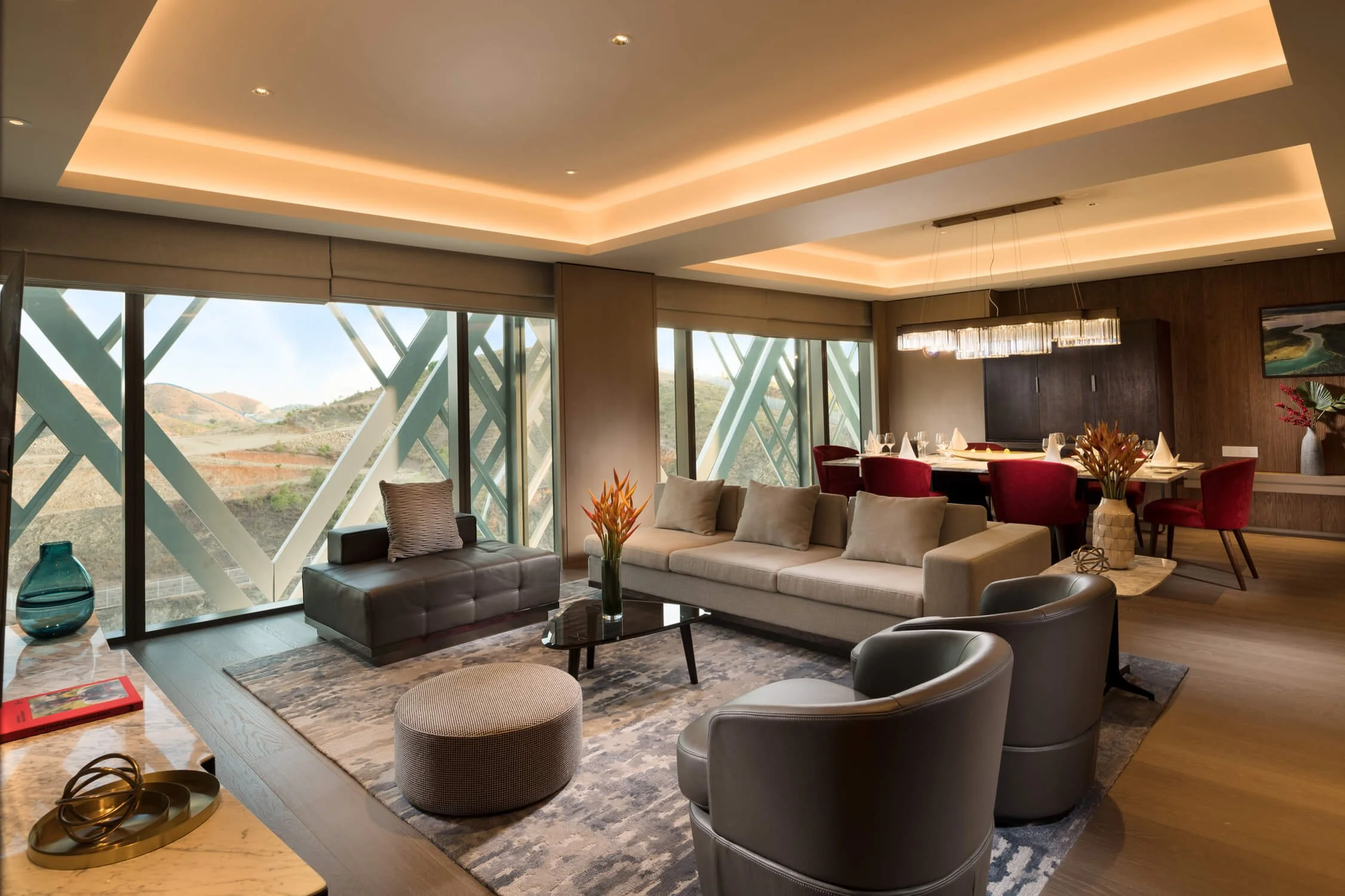 Modern living room with large windows, beige sofa, black armchair, round ottoman, and dining area with red chairs, with mountain view outside.