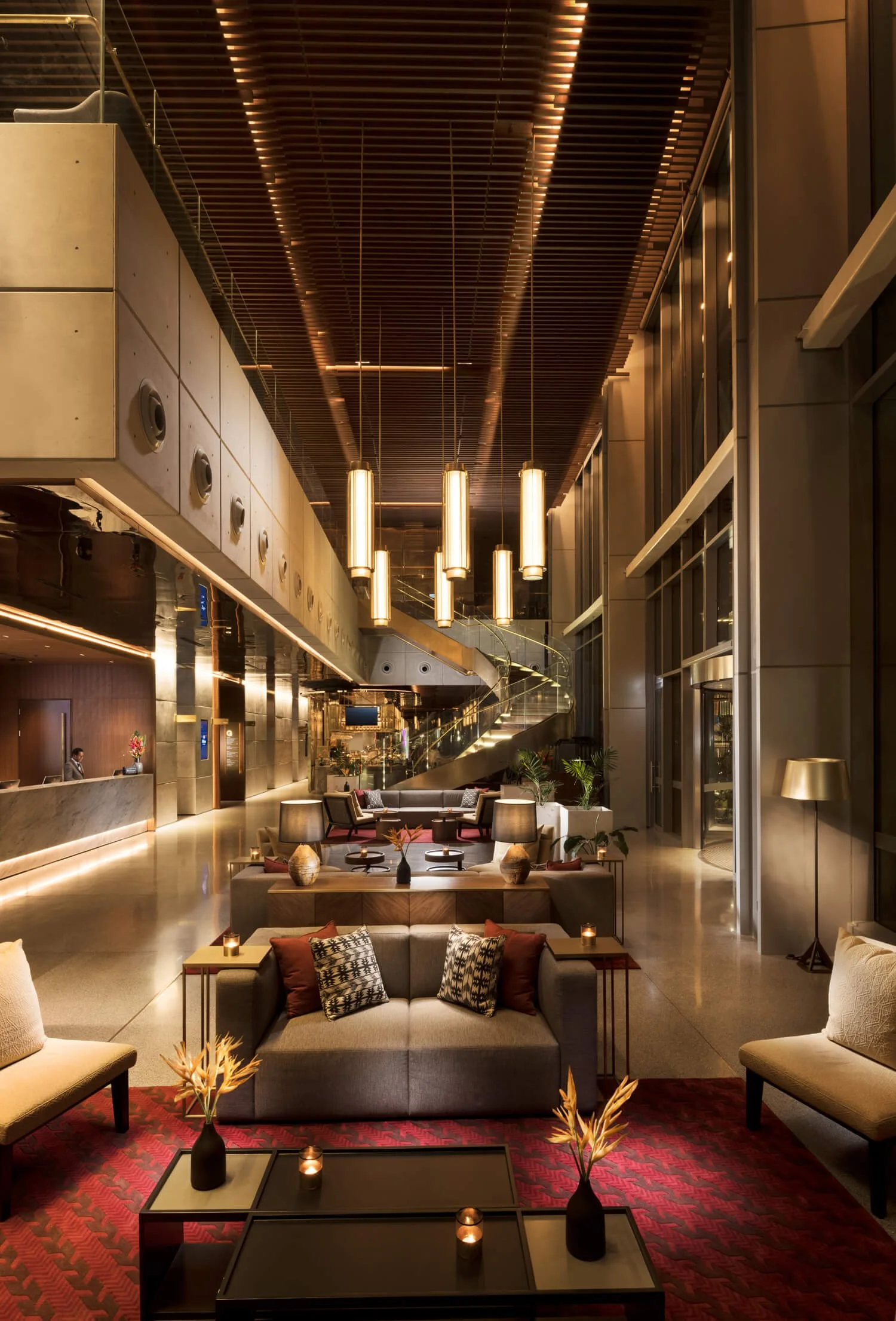 Luxurious hotel lobby with modern decor, plush seating, a fireplace, decorative vases and plants, a winding staircase, and warm lighting.