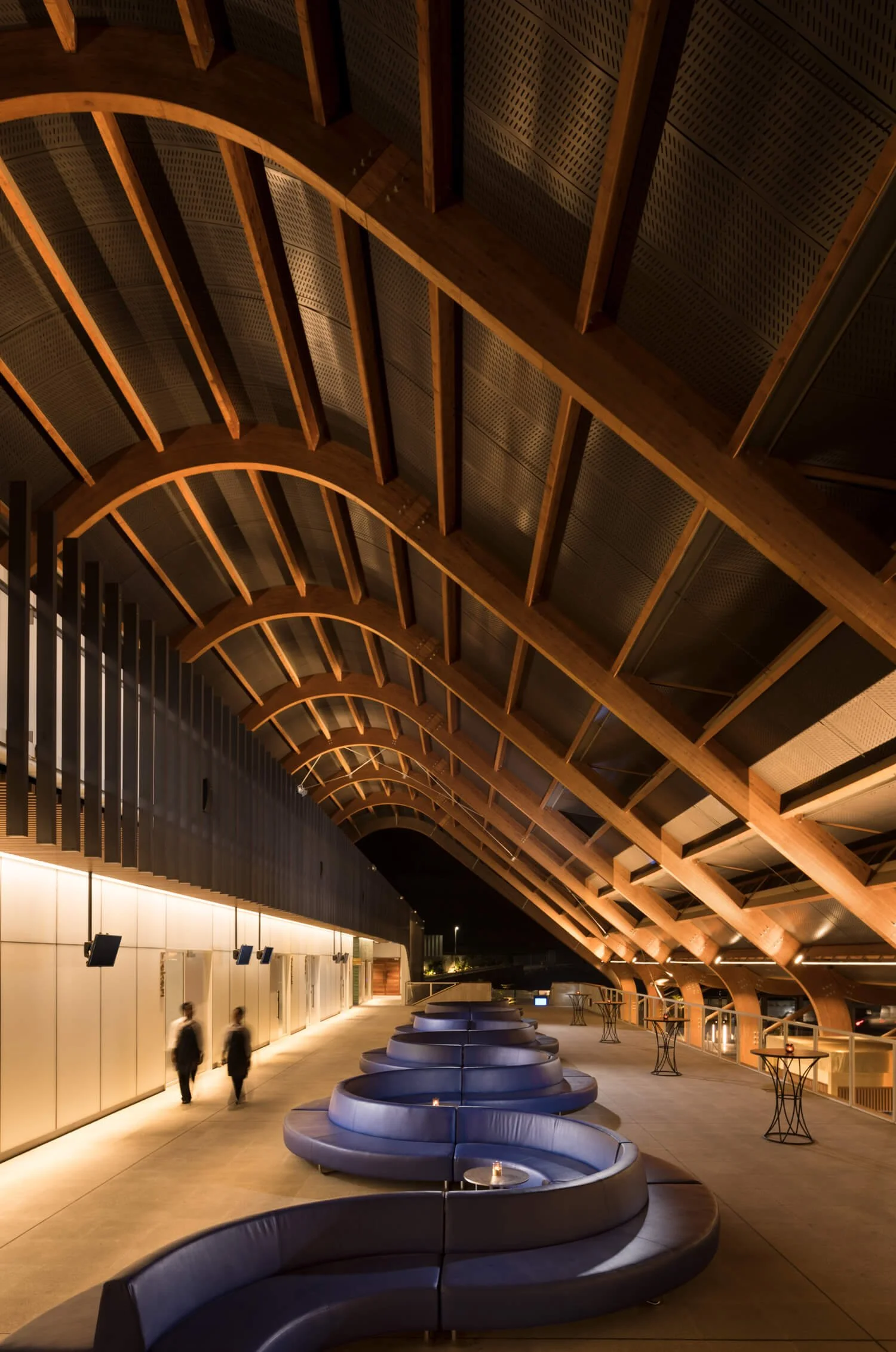 Interior of a modern building with a curved wooden ceiling, blue semi-circular seating, small tables with lit candles, and two people walking by at night.