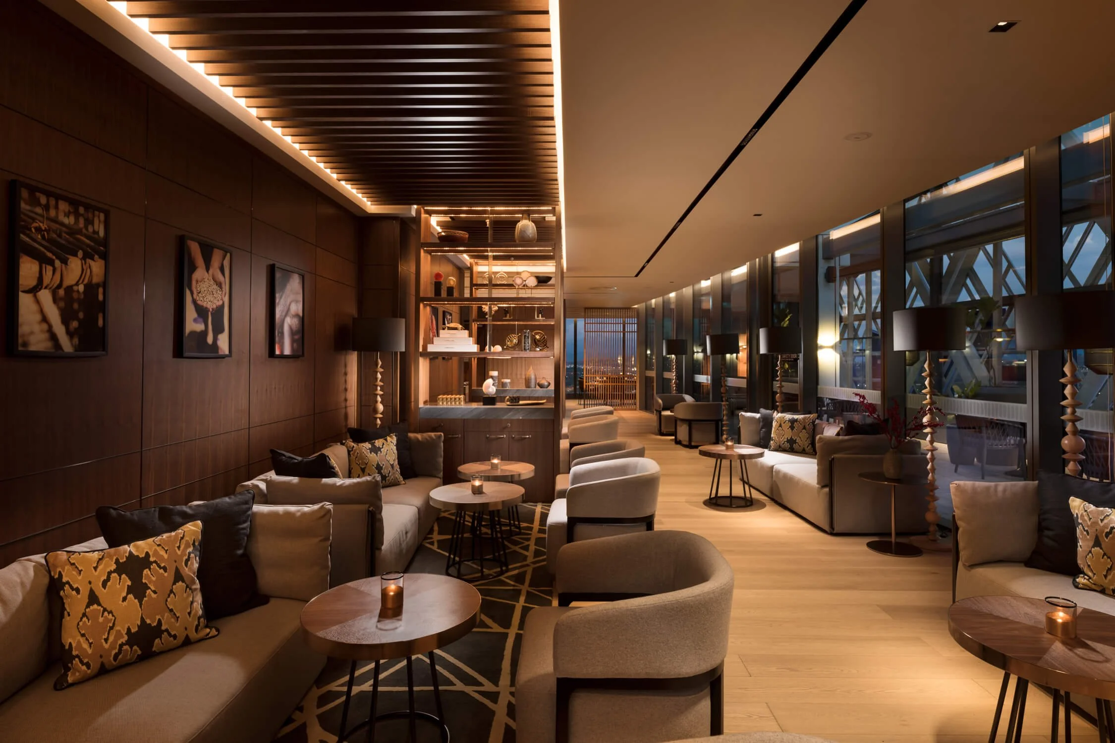 A luxurious lounge with beige sofas, armchairs, and wooden tables with candles, set against dark wood-paneled walls and large floor-to-ceiling windows showing an outdoor area with a bridge structure, modern black lamps, decorative pillows, and shelving with books and ornaments.