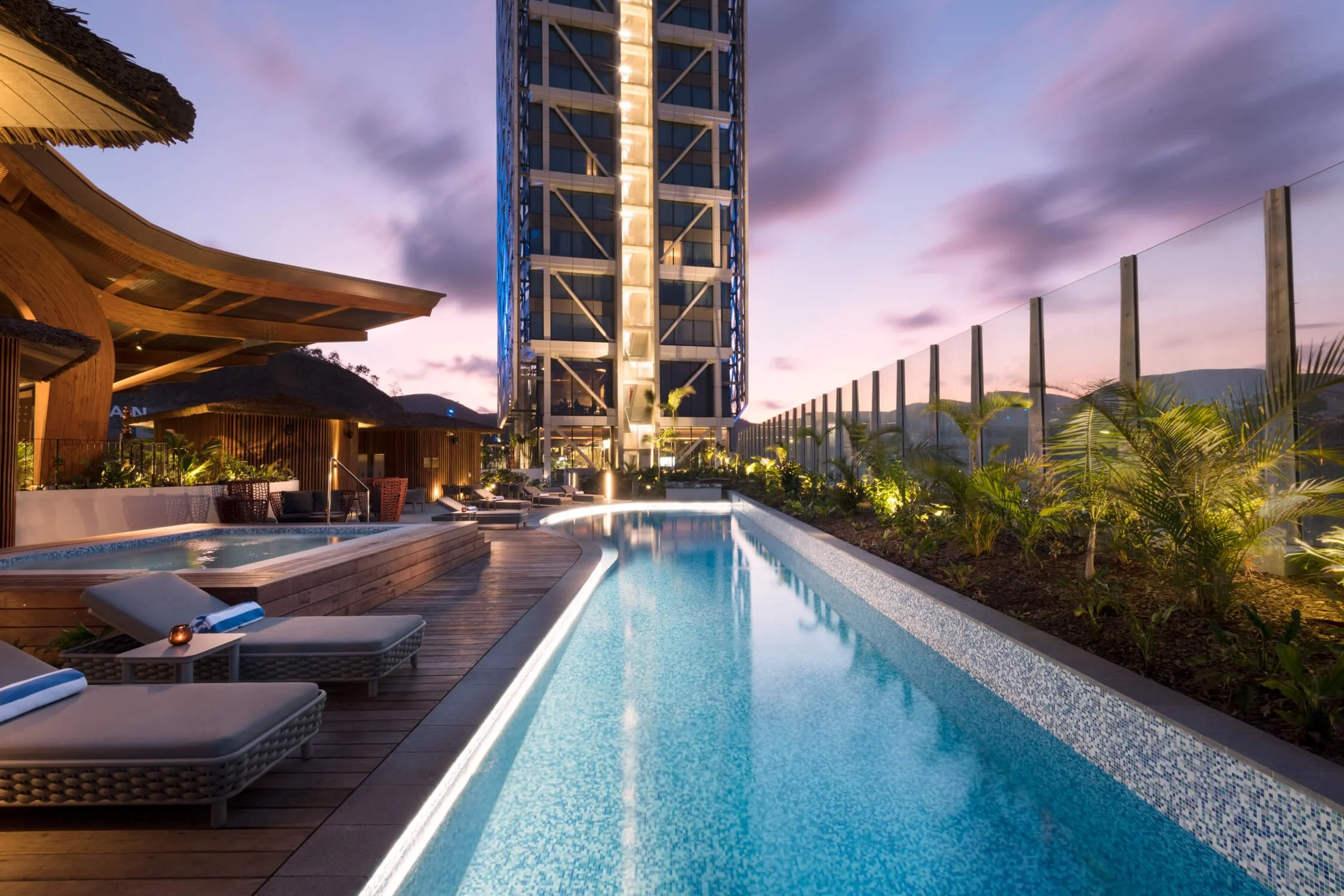 Luxury rooftop pool area at sunset with lounge chairs, a hot tub, modern building in background, and cityscape mountains in distance