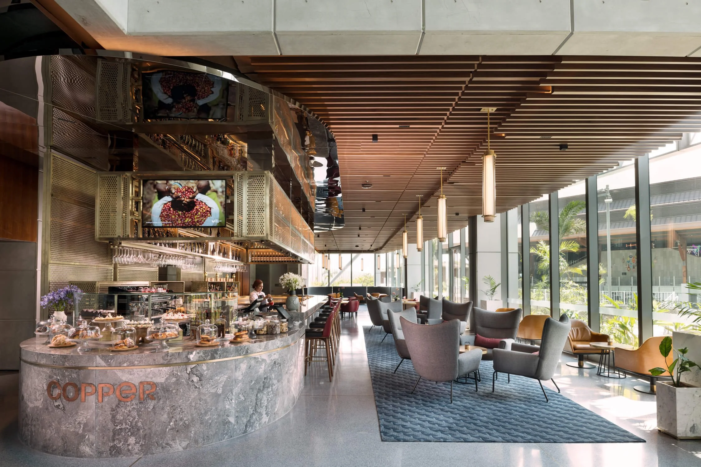 Modern cafe with marble counter, bar stools, and seating area with armchairs and small tables, large glass windows with outdoor greenery, and wooden ceiling panels.