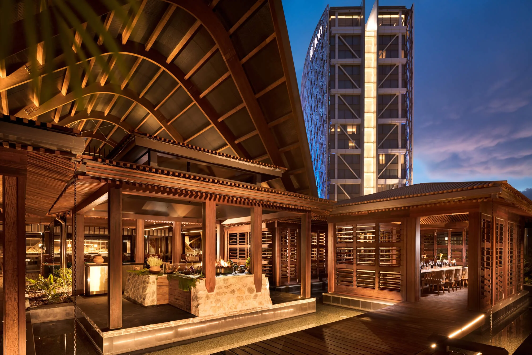 Upscale outdoor patio area with wooden structure and seating, illuminated by warm lighting, featuring a traditional Japanese or Asian-inspired architectural style, with a modern high-rise building in the background during evening or night.