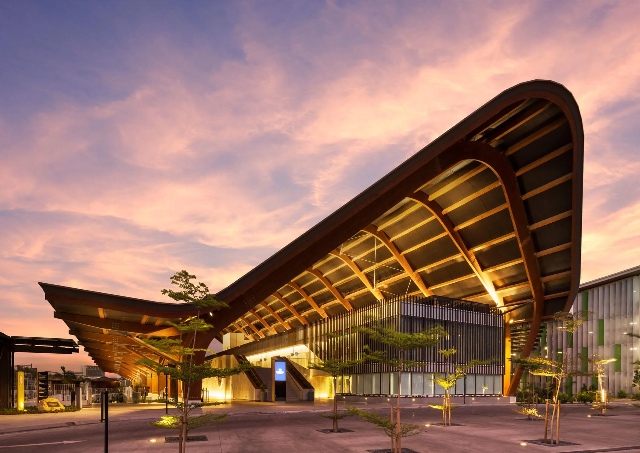 Modern building with curved roof structure, lit from below, against a pink and purple sunset sky, surrounded by small trees and parking lot.
