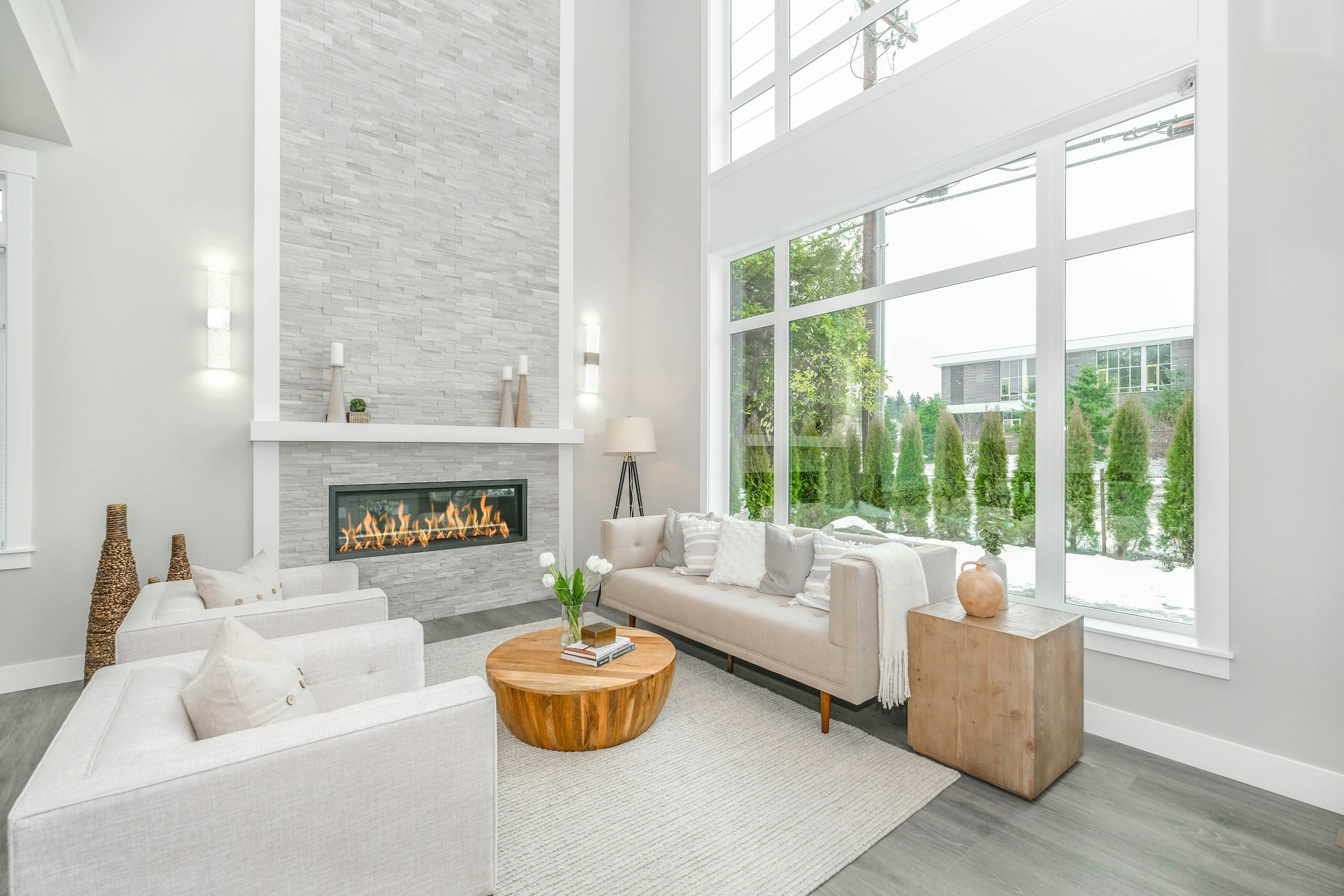 Bright living room with large windows showing greenery outside, white walls, gray wood floor, modern fireplace with lit flames, white sofas, wooden coffee table with flowers, throw pillows, minimalist decor.