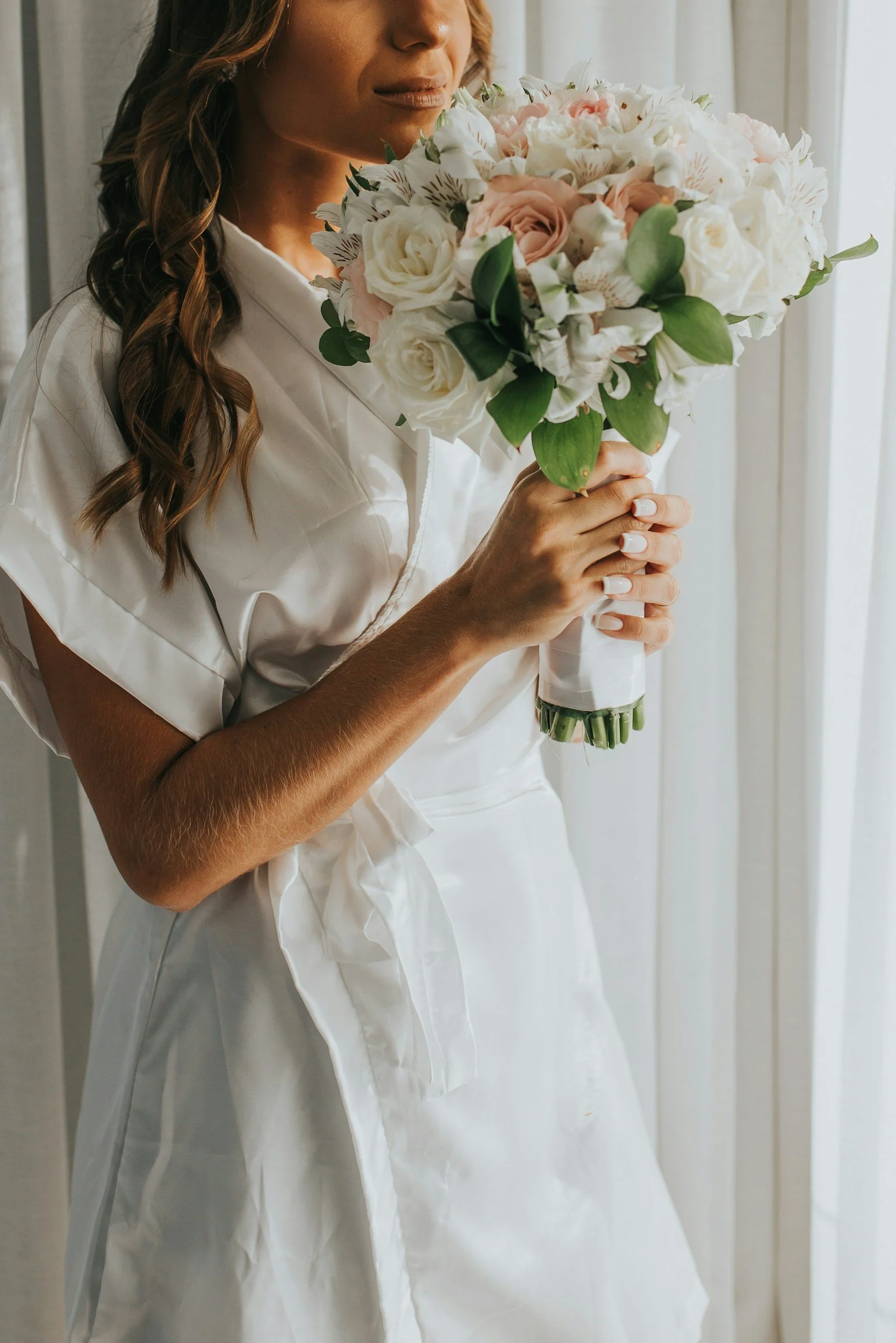 A woman in a white dress holding a bouquet of white and blush pink roses, white flowers, and green leaves, standing near a window.