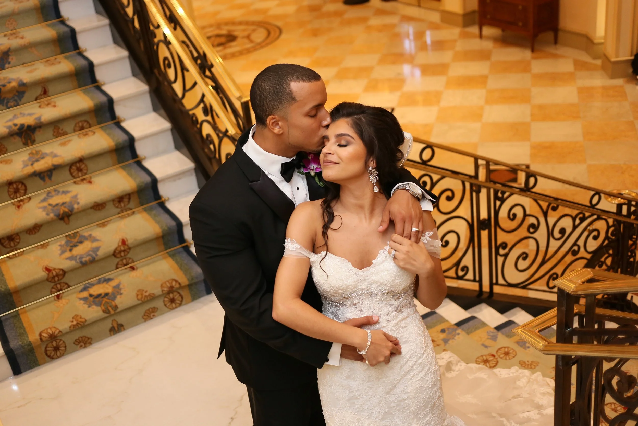 A newlywed couple sharing an intimate moment on a staircase with a decorative carpet, elegant black railing, and polished tile floor in an upscale venue.