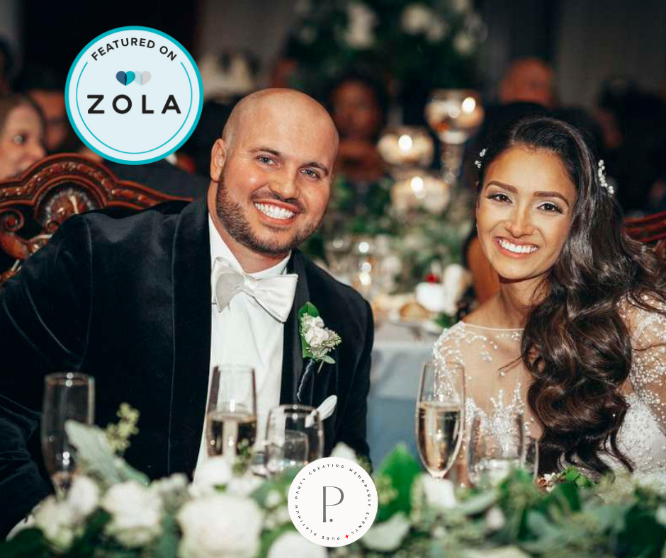 A happy bride and groom sitting at their wedding reception, smiling at the camera, dressed in elegant wedding attire, with a decorated table and guests in the background.