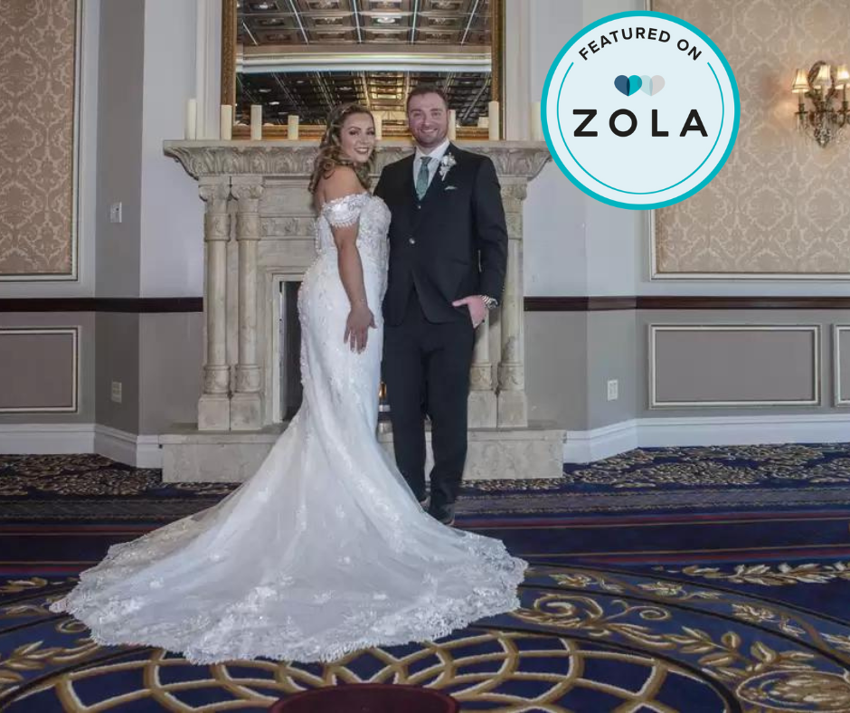 Bride and groom in wedding attire standing in front of a fireplace with an ornate mirror, in a decorated wedding venue, with a Zola feature badge in the top right corner.