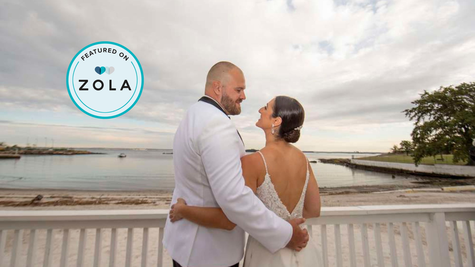A newlywed couple embracing on a beach with water, boats, and a cloudy sky in the background.
