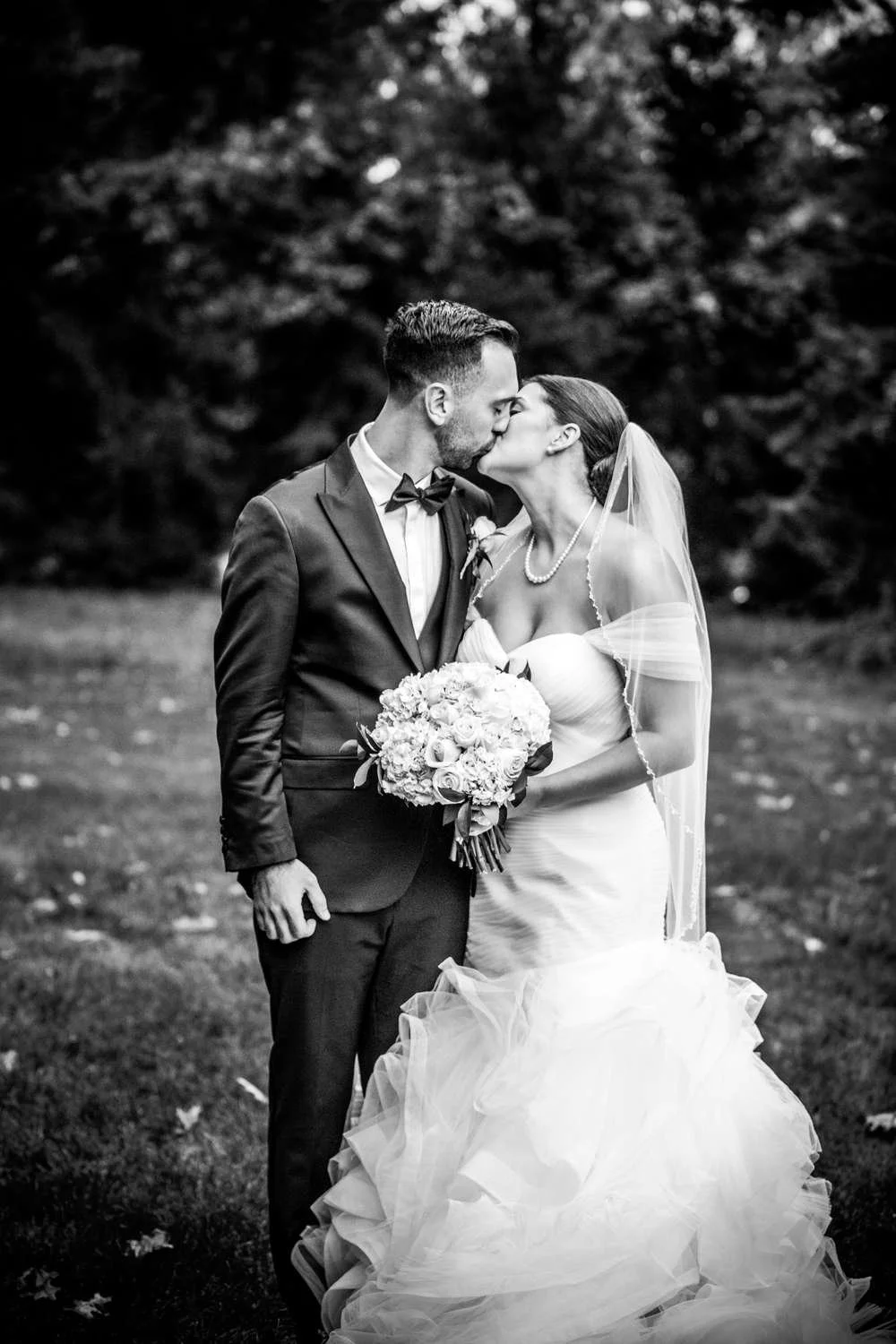 Black and white photo of a bride and groom kissing outdoors, with the bride holding a bouquet of flowers, both dressed in wedding attire.