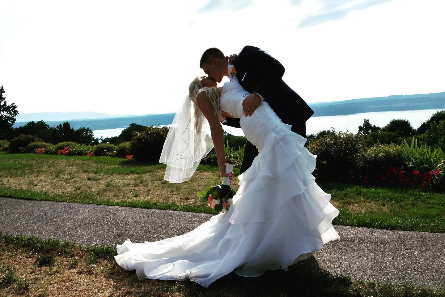 A bride in a white wedding dress and veil leans back while a groom in a black suit kisses her, outdoors near a lake with green trees and bushes in the background.