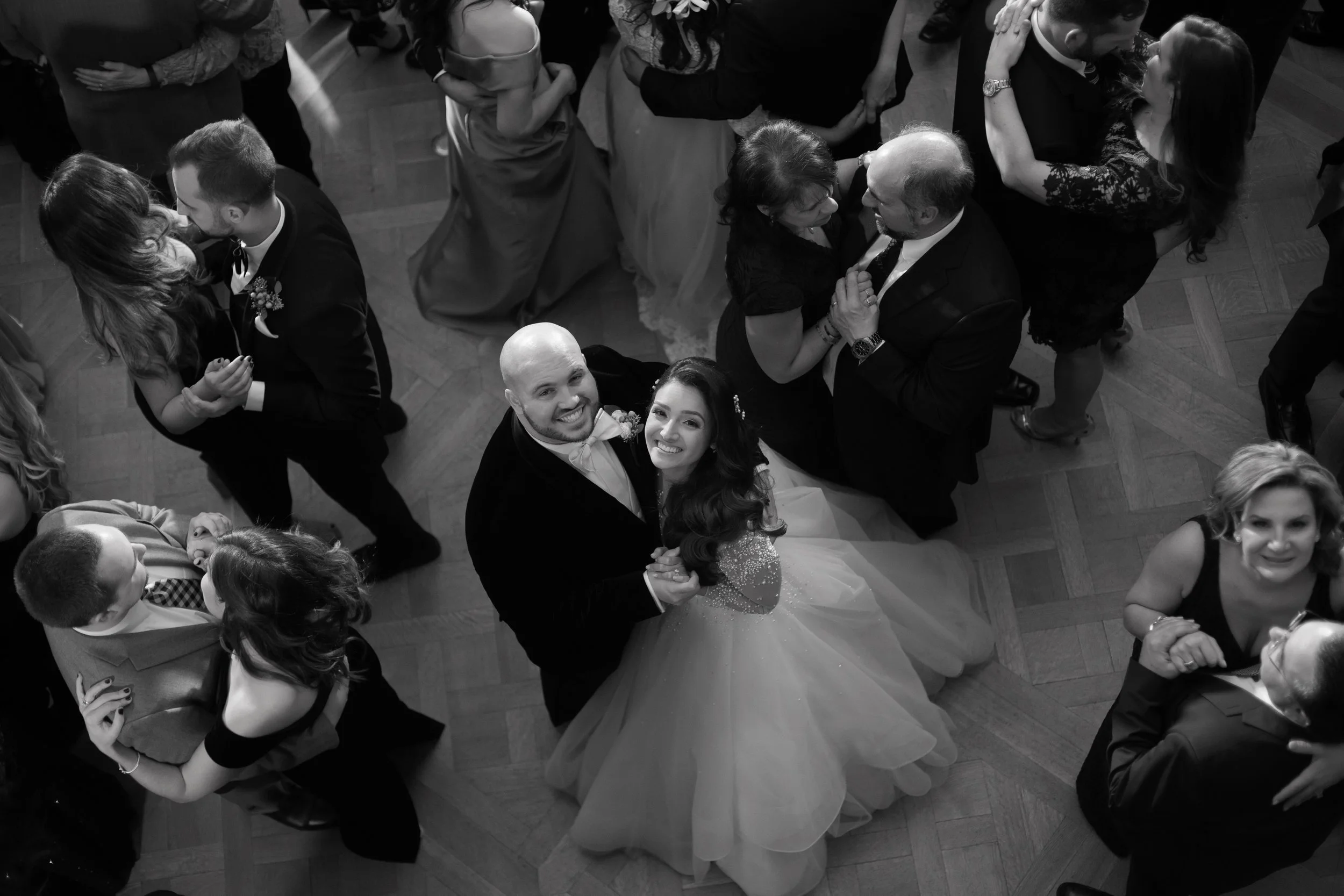 Black and white photo of a wedding dance floor with multiple couples dancing, including a bride and groom holding hands and smiling at the camera.