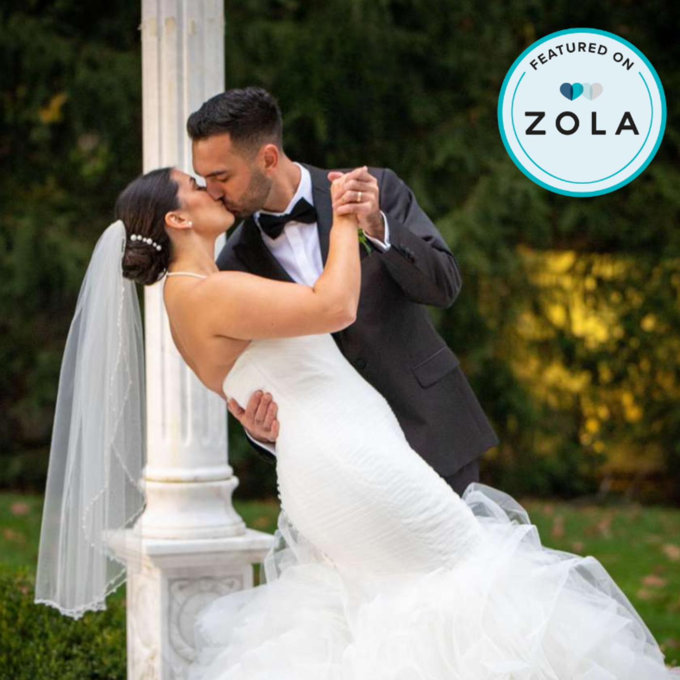 A bride and groom share a kiss during their wedding ceremony outdoors, with the groom dipping the bride as she wears a strapless white wedding gown and a veil, and the bride wearing pearl earrings and a hair accessory.