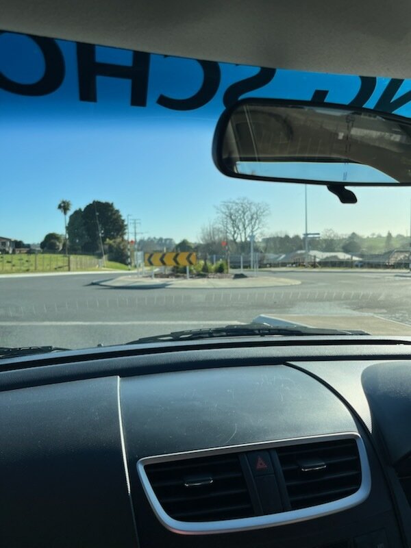 View from inside a car at a roundabout with a yellow and blackChevron directional sign and trees in the background.