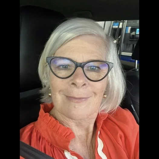 An older woman with short white hair and glasses, smiling in a car, wearing a red top.