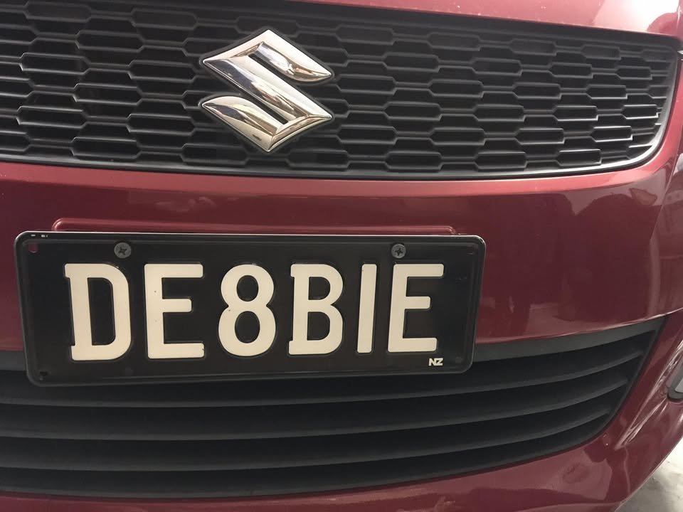 Close-up of a red Suzuki car with a New Zealand license plate reading 'DE8BIE'.