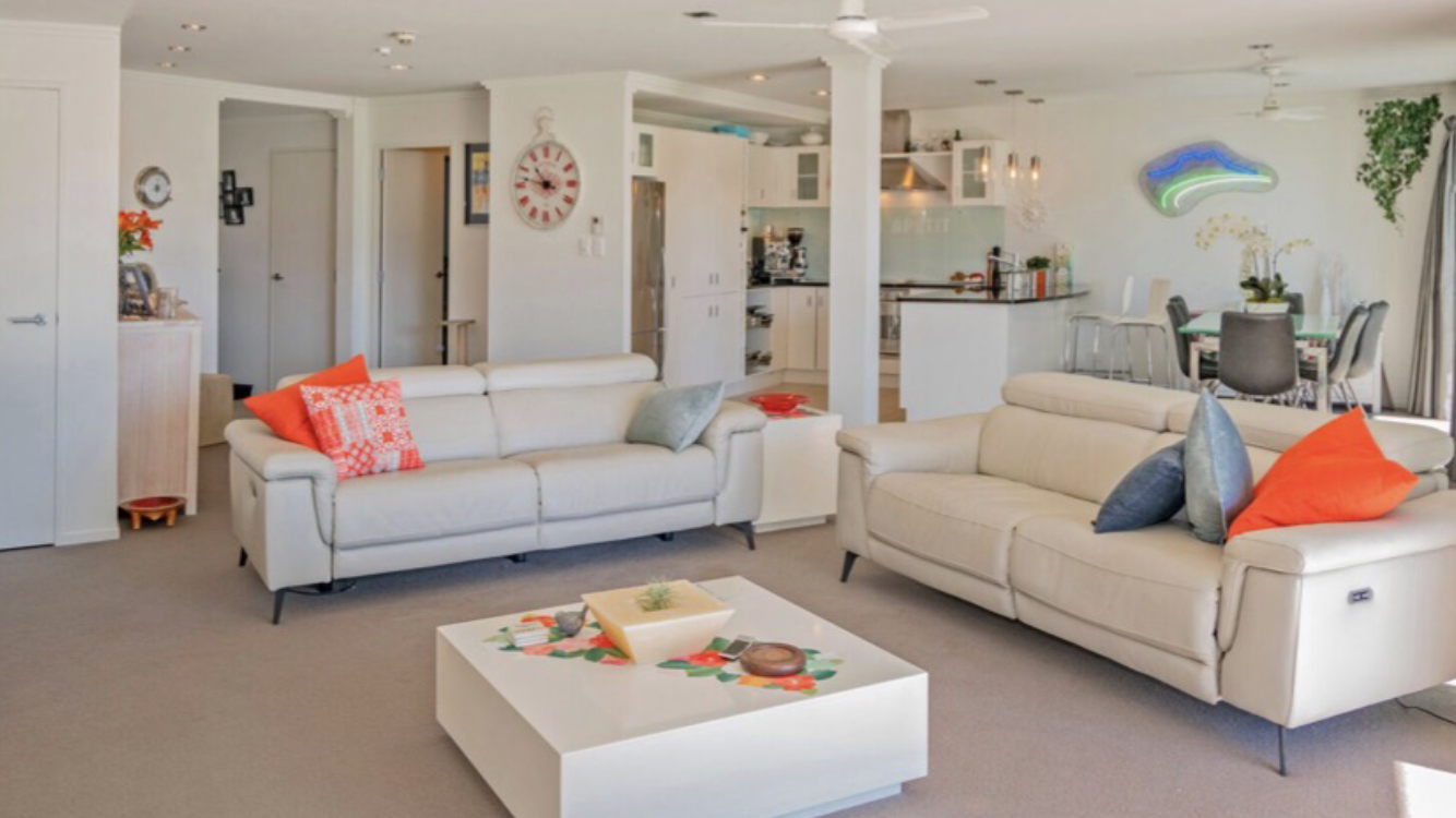 Living room with two white sofas, colorful throw pillows, a white coffee table, and open kitchen and dining area in background.