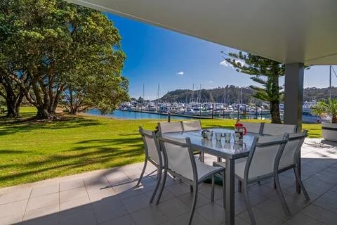Outdoor patio with dining table and six chairs, overlooking a marina with boats and sailboats, under a covered area with green lawn and trees in the background.
