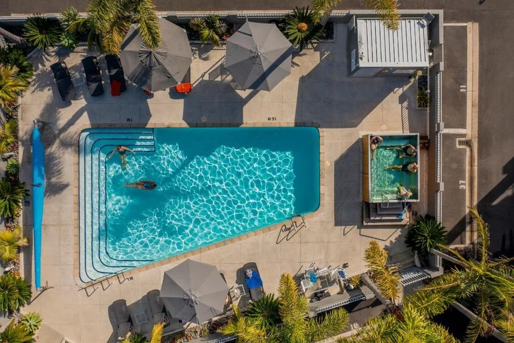 Overhead view of a swimming pool with a person swimming and several people in a hot tub, surrounded by lounge chairs, umbrellas, and palm trees.