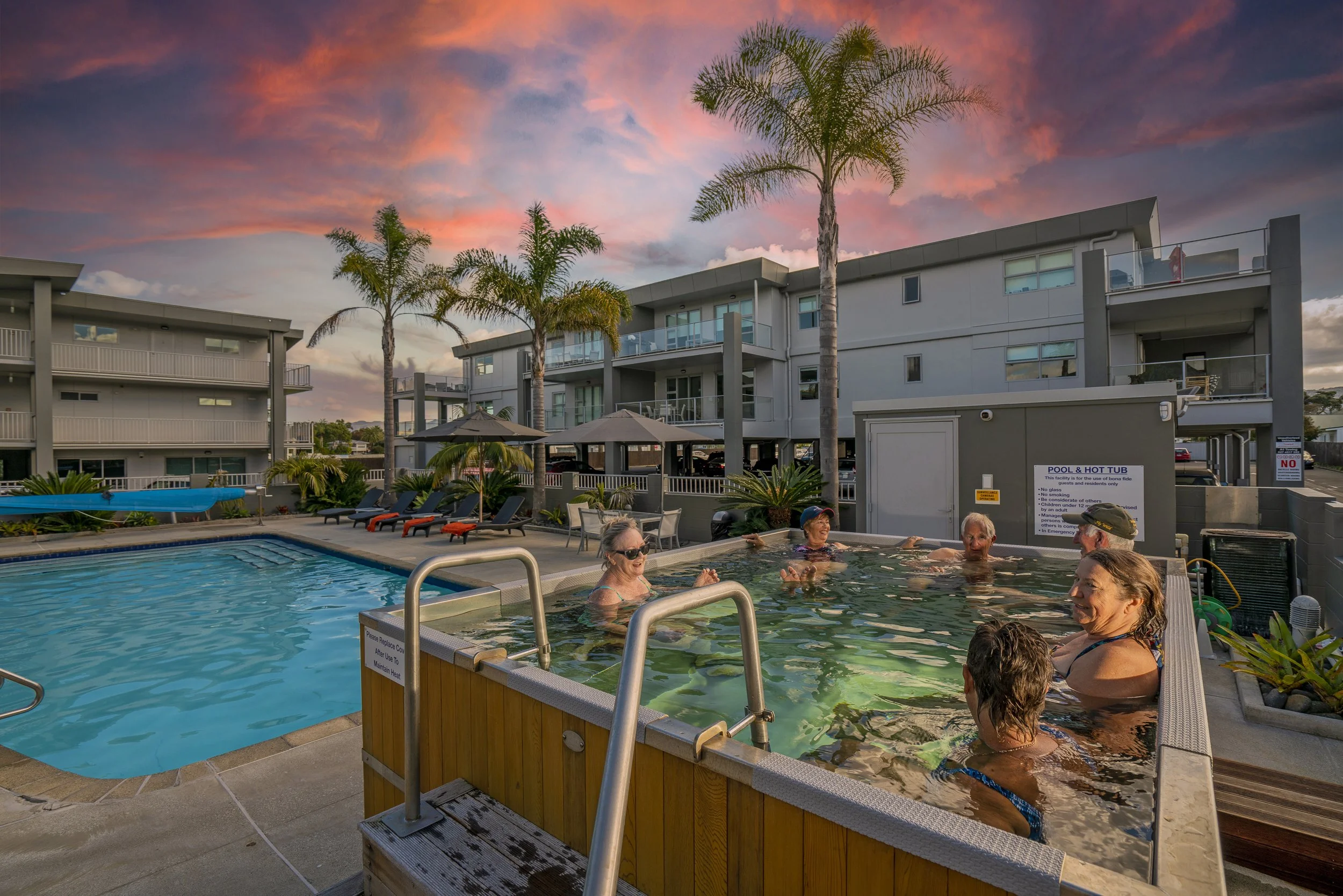 A group of people enjoying a hot tub and a swimming pool at sunset in an apartment complex.