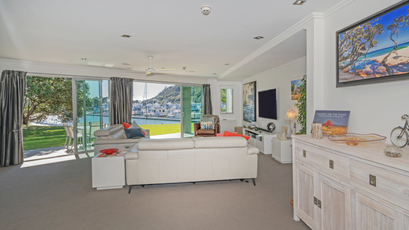 Living room with white sofa, brown armchair, and a flat-screen TV on the white wall facing large sliding glass doors leading to a patio and a view of a marina with boats and a hillside.
