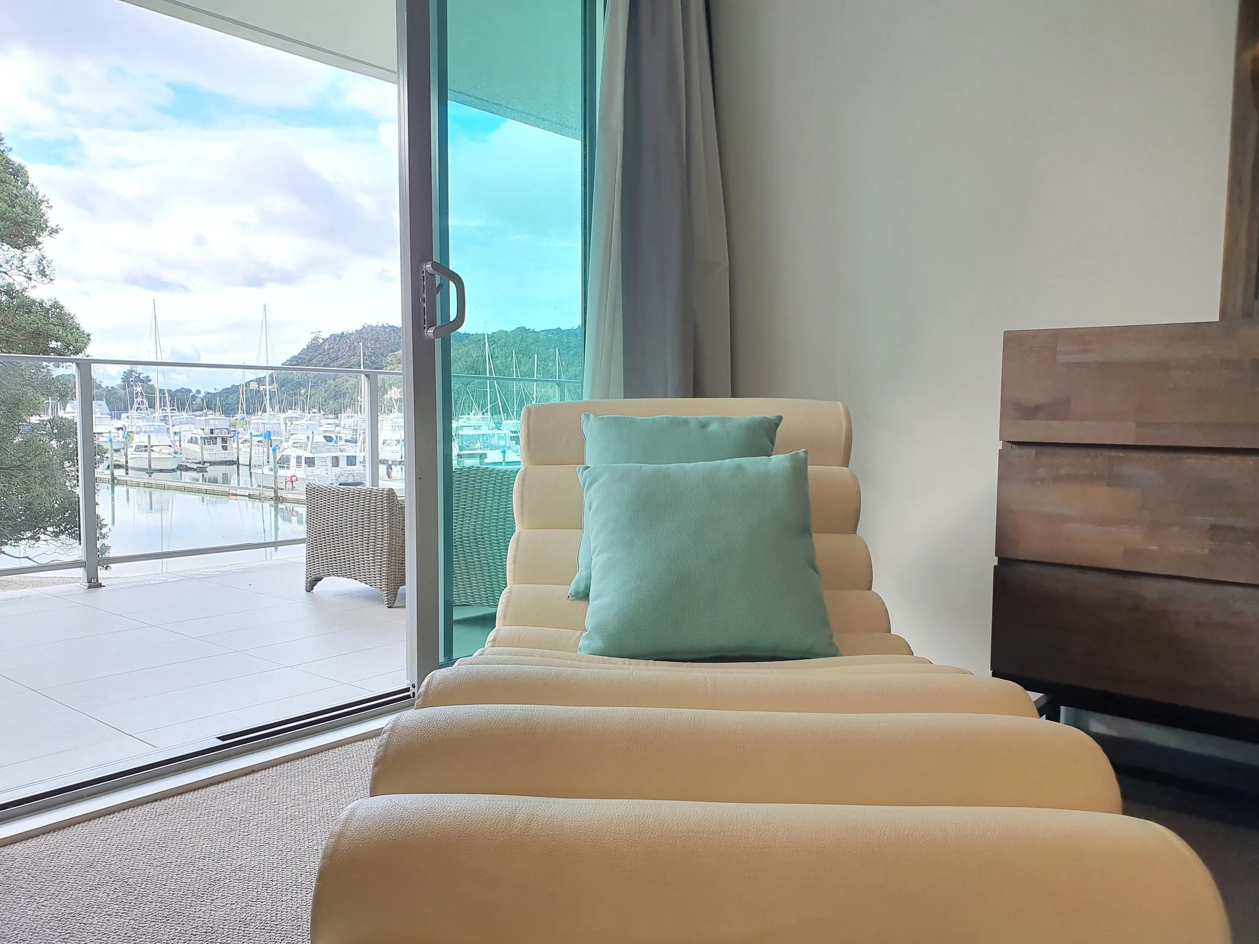 Living room with a cream-colored lounge chair with two teal pillows, adjacent to a wooden dresser, and a large sliding glass door leading to a balcony with a view of boats at a marina and a hill in the background.