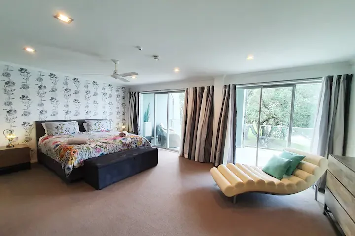 A spacious bedroom with a king-sized bed with floral bedding and pillows, a nightstand with a lamp, a patterned wallpaper behind the bed, a ceiling fan, large sliding glass doors with curtains leading to a balcony, and a modern cream-colored lounge c