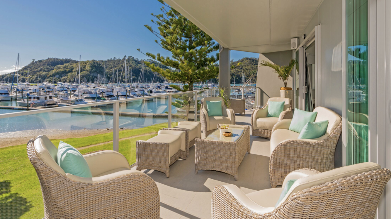 Balcony with wicker furniture and cushions overlooking a marina with boats and sailboats, surrounded by green hills under a clear blue sky.