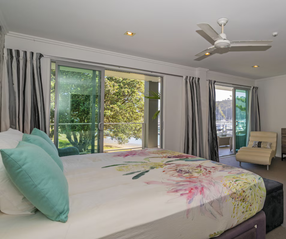 A bedroom with a large bed, turquoise pillows, floral bedspread, and sliding glass doors leading to a balcony with trees and a view of a body of water.