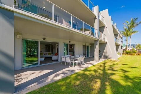 Modern apartment building with large glass doors and balcony, grassy yard with trees, clear blue sky.